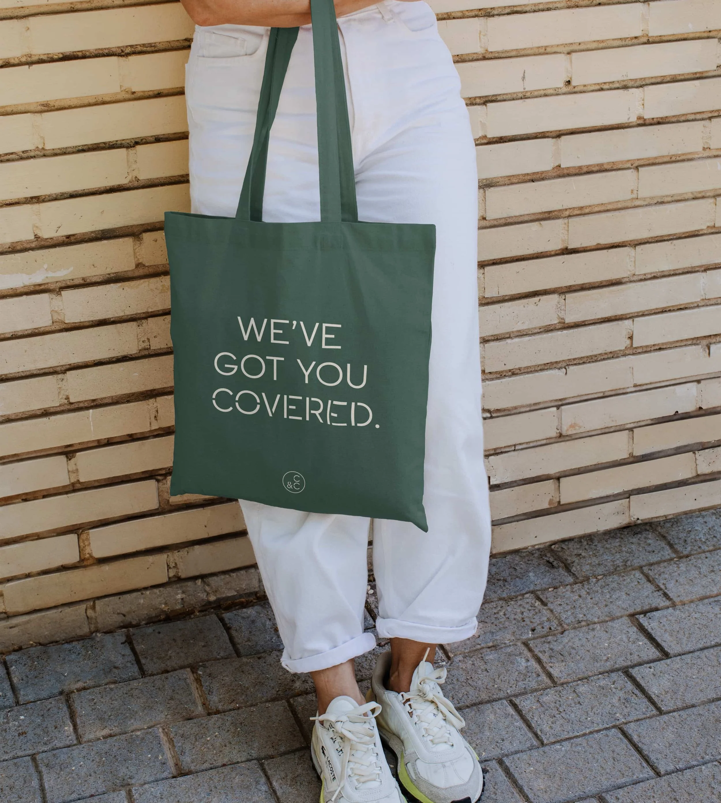 Person standing against a brick wall, wearing white pants and white sneakers, holding a green tote bag with the words 'WE'VE GOT YOU COVERED' printed on it.
