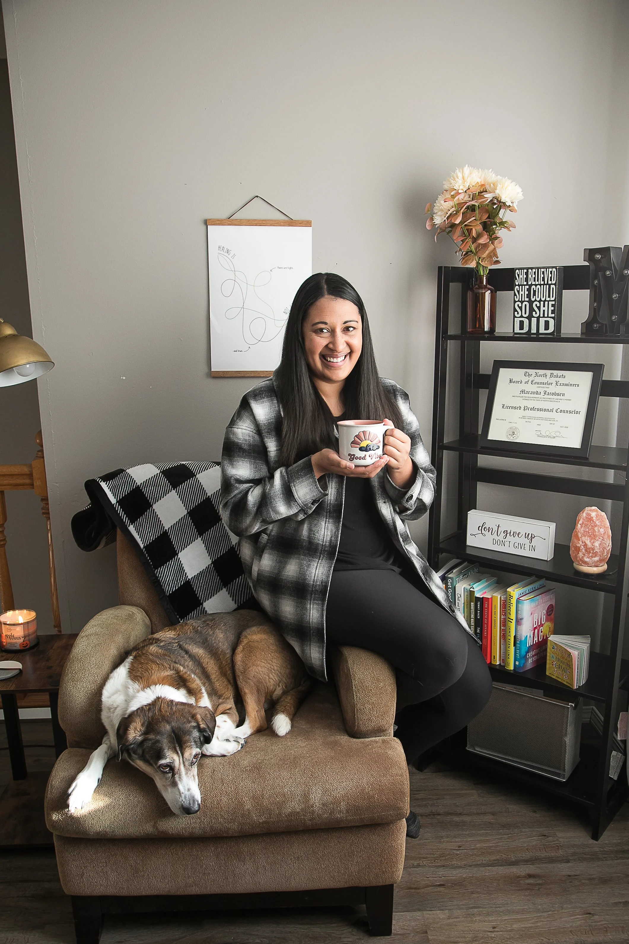 A woman smiling and holding a mug, sitting on a brown armchair with a large dog lying on the chair in front of her. The setting is a cozy living room with a bookshelf, framed certificate, and decorative items including a vase with flowers and motivational signs on the shelves.