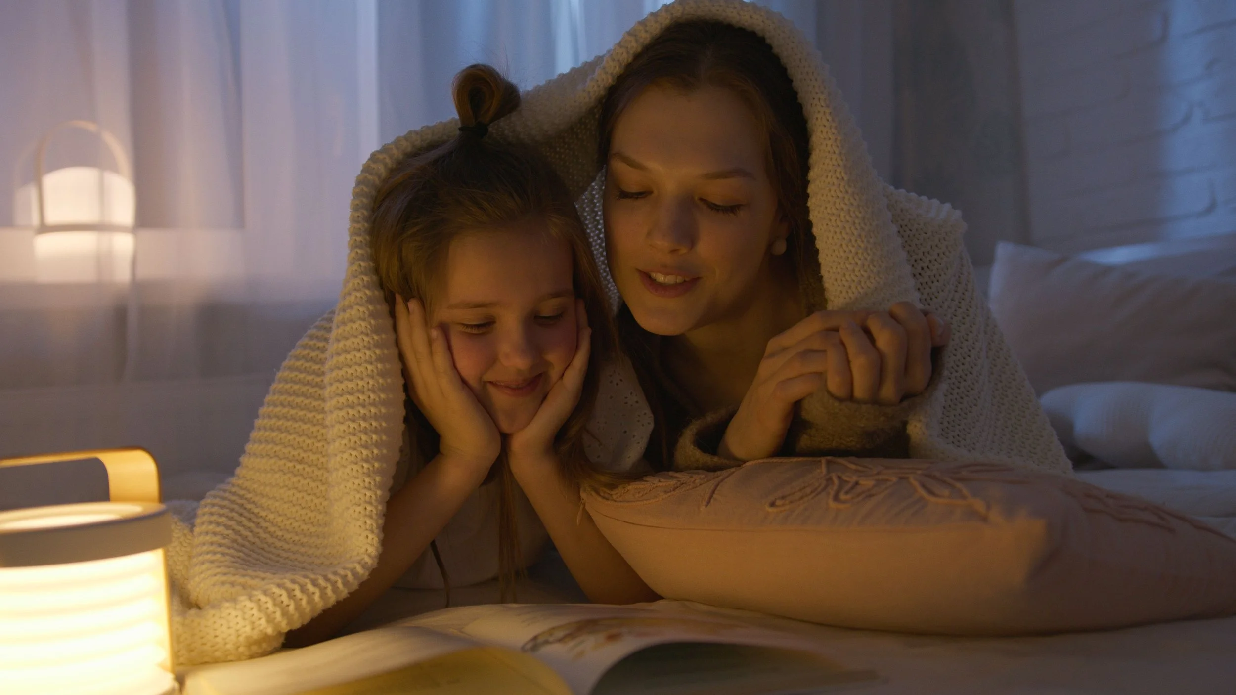 A woman and a girl are under a blanket on a bed, smiling and enjoying a cozy moment together in a softly lit room.
