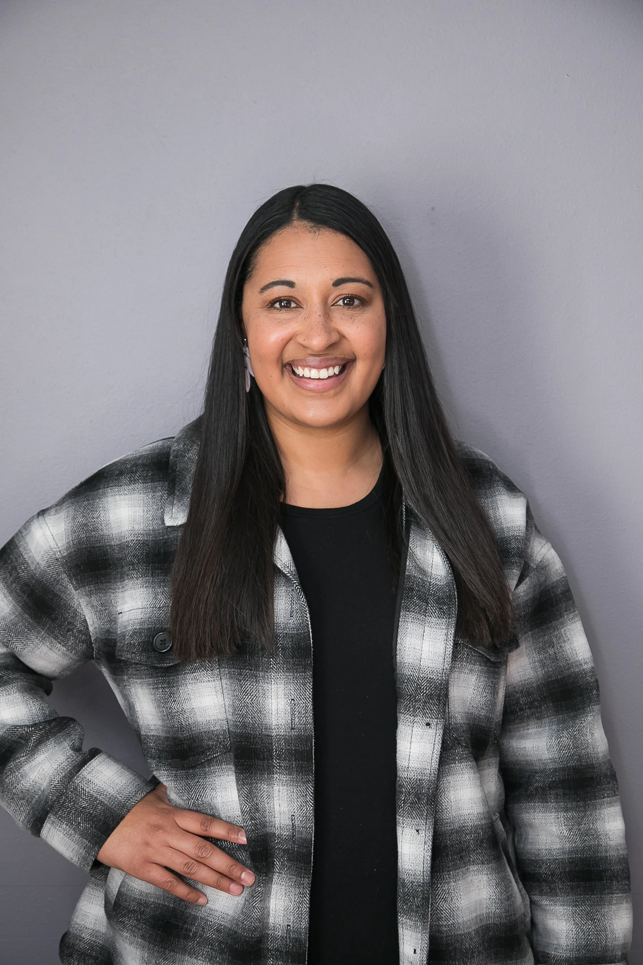 A woman with long black hair, wearing a black shirt and a black-and-white plaid jacket, standing against a plain gray background, smiling with her hand on her hip.