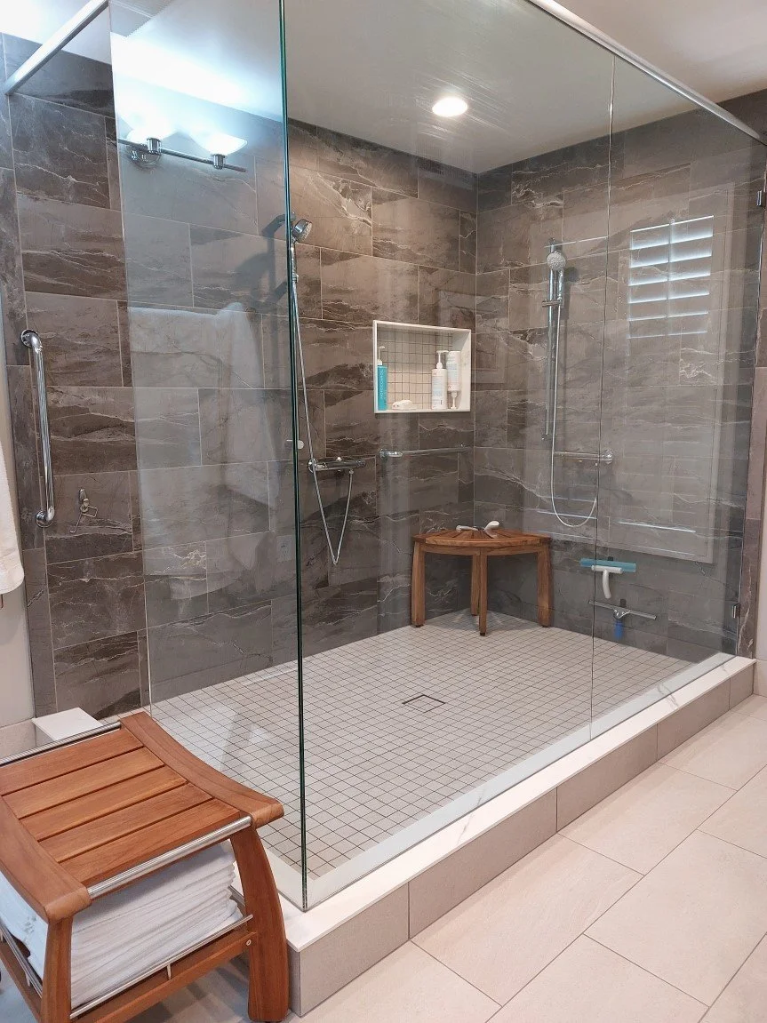 Modern glass-enclosed shower with brown tile walls, small white tiled floor, wooden bench, and built-in shelf with toiletries.