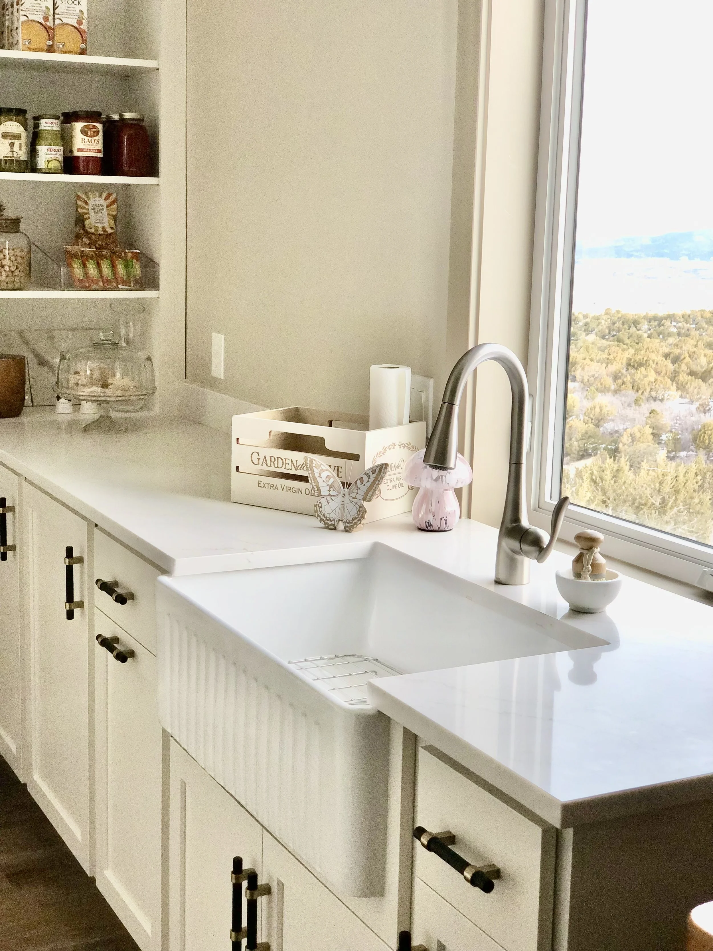 A modern kitchen with white cabinets, a white farmhouse sink, a silver faucet, and a large window overlooking a landscape. On the counter are a tissue roll, a soap dispenser, a white bowl with a utensil, and decorative items including a butterfly and a pink glass flower. Shelves on the wall hold jars and snacks.