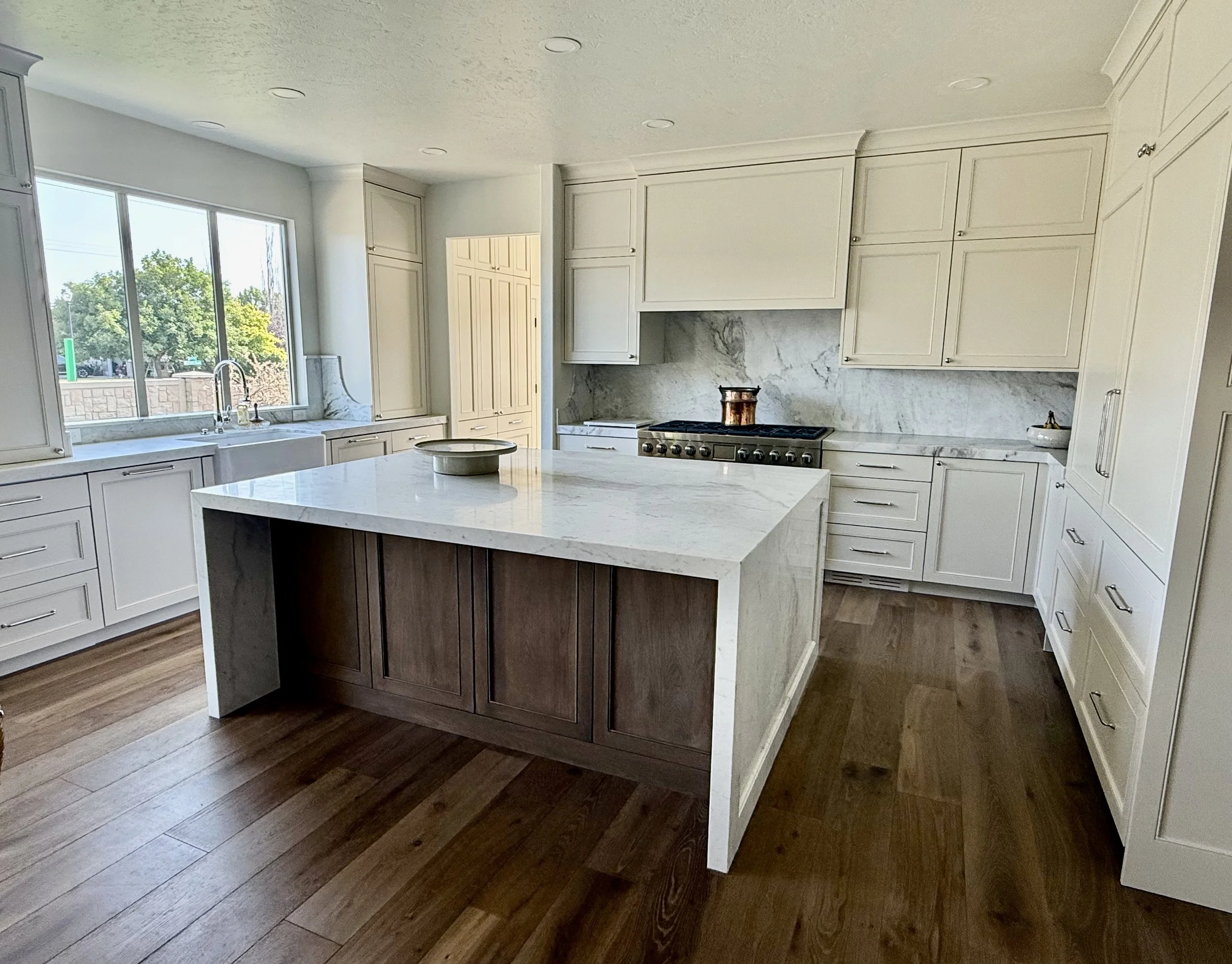 Modern kitchen with white cabinets, marble countertops, and a large kitchen island with a wood bottom. Window above the sink shows trees outside.