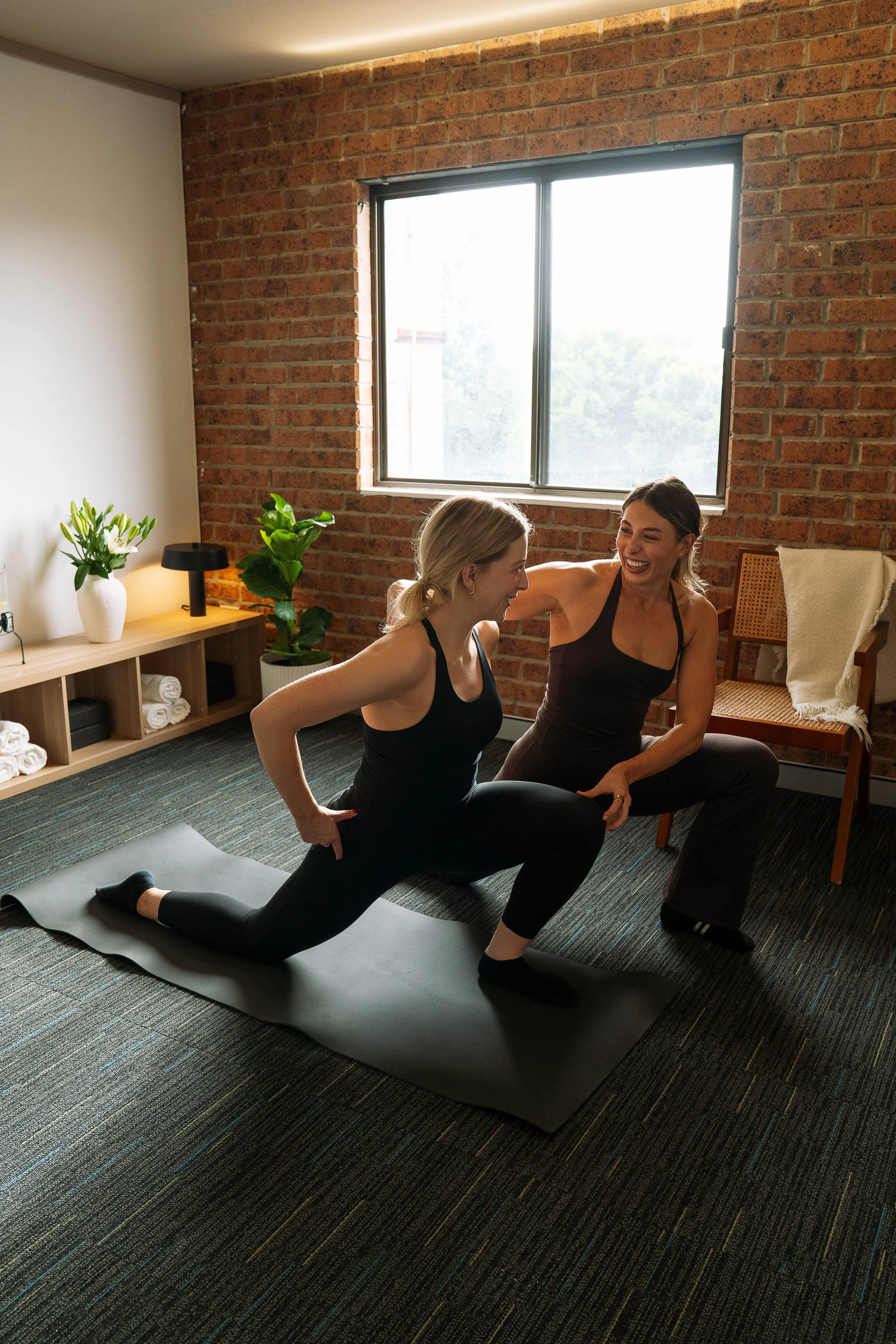 Two women with dark hair are practicing yoga together in a cozy room with an exposed brick wall. One woman is in a lunge position on a yoga mat, and the other woman is kneeling and smiling, helping or encouraging her.