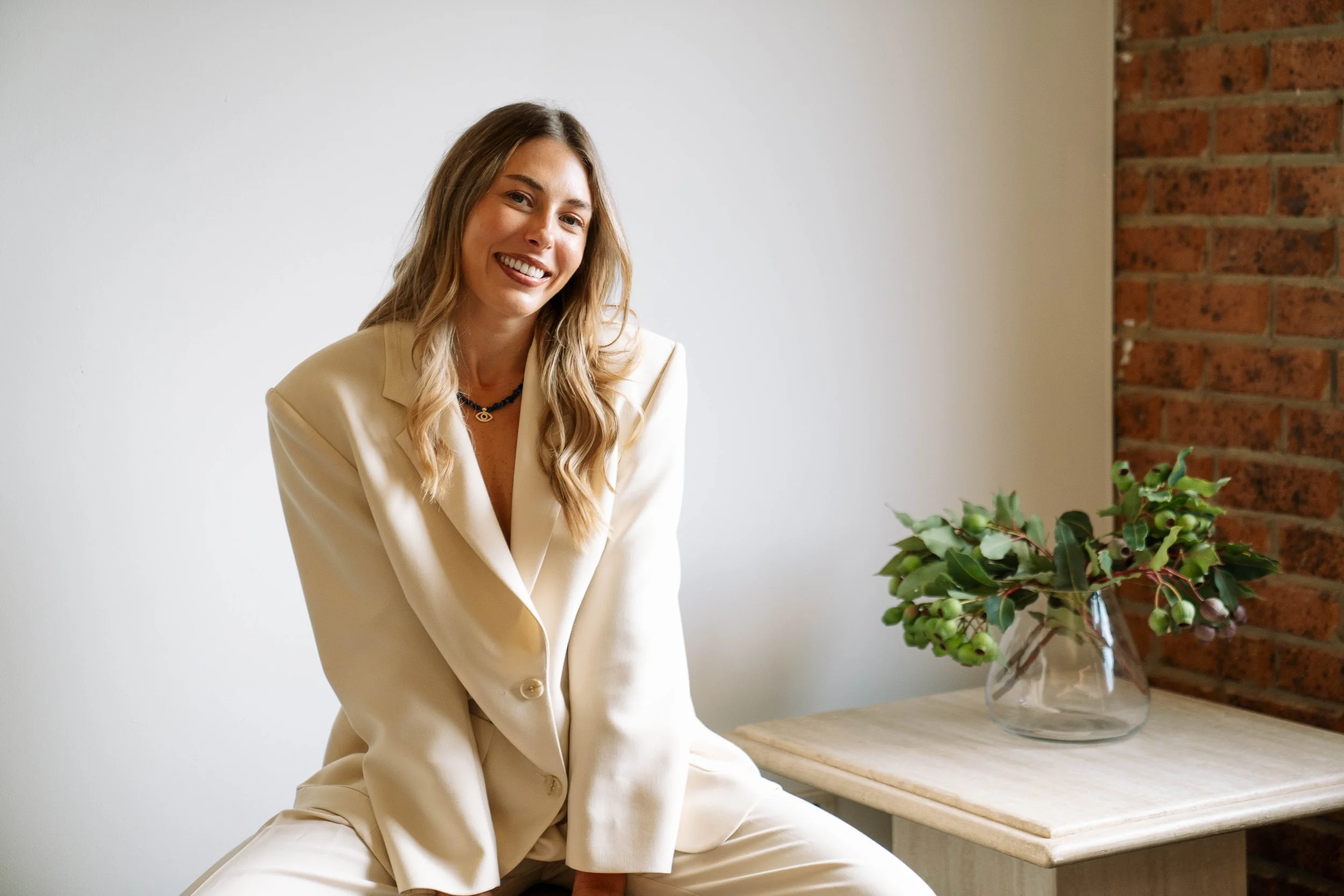 A woman in a white suit sitting by a table with a large glass vase holding green and purple plants, in front of a white wall and a red brick wall.