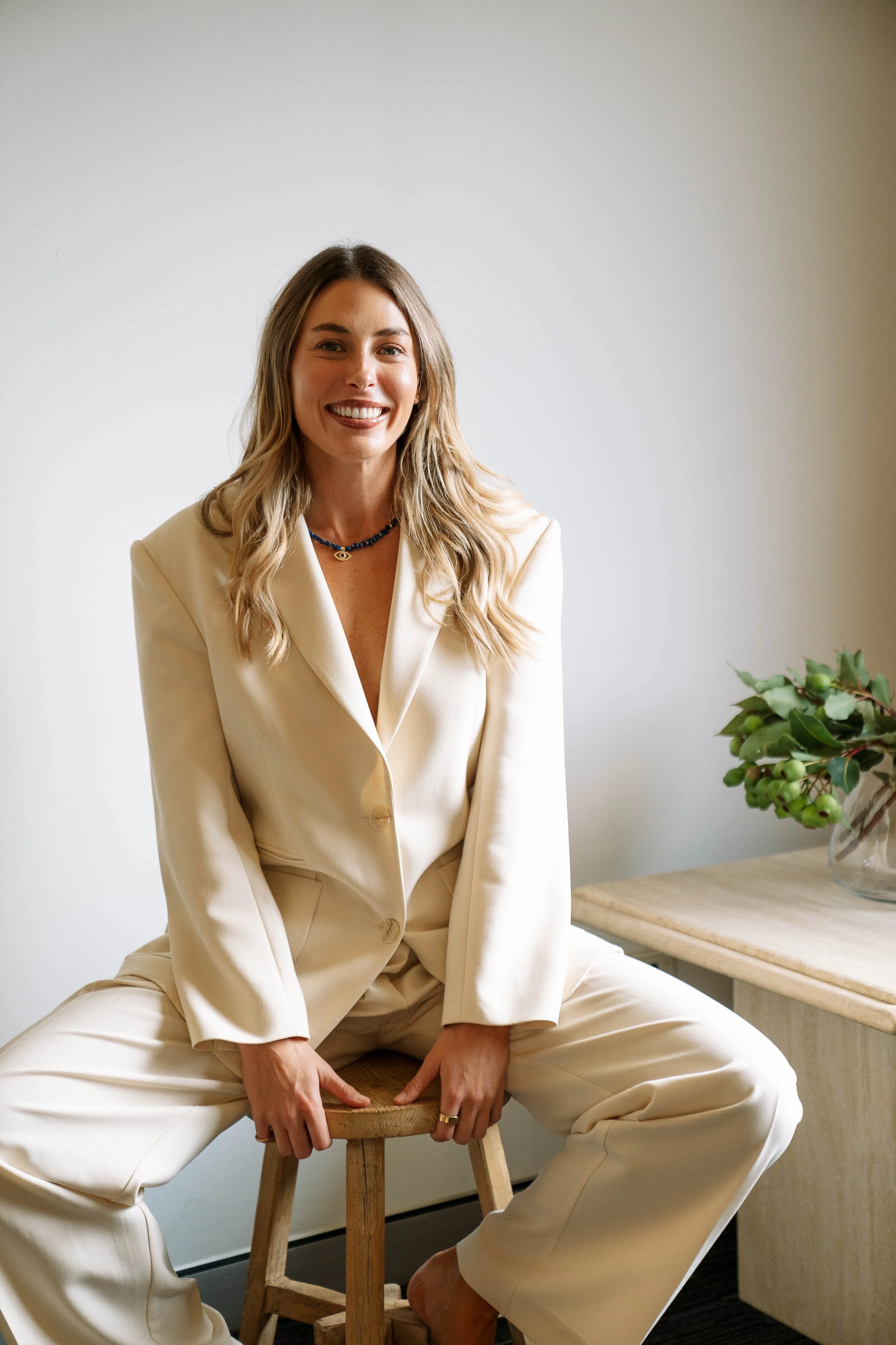 A woman with long wavy blonde hair wearing a cream-colored suit is sitting on a wooden stool, smiling at the camera. She is in a room with a white wall, a wooden table, and a potted plant on the table.