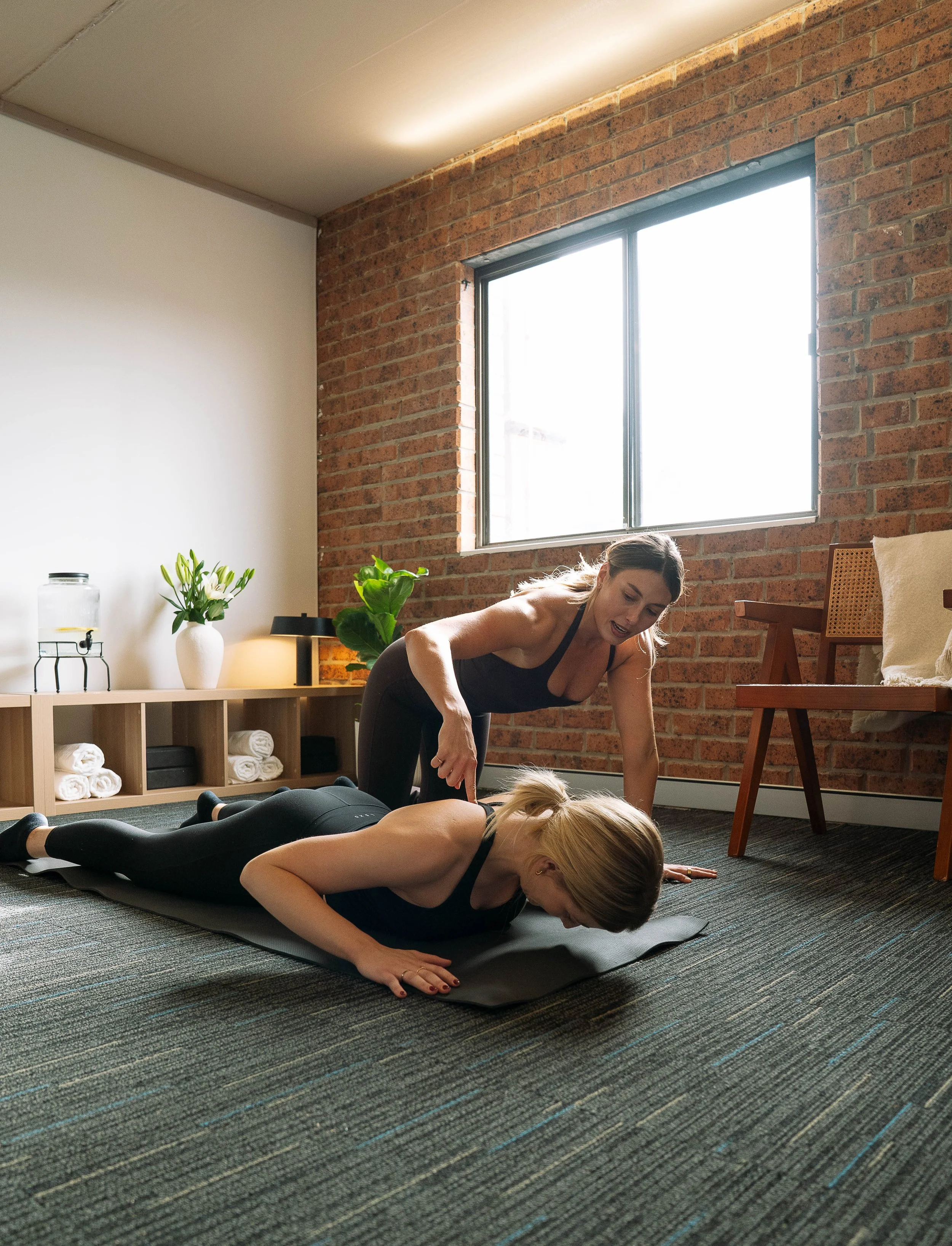 Two women doing yoga in a brick-walled apartment, with one woman assisting the other in a plank position on a black yoga mat.