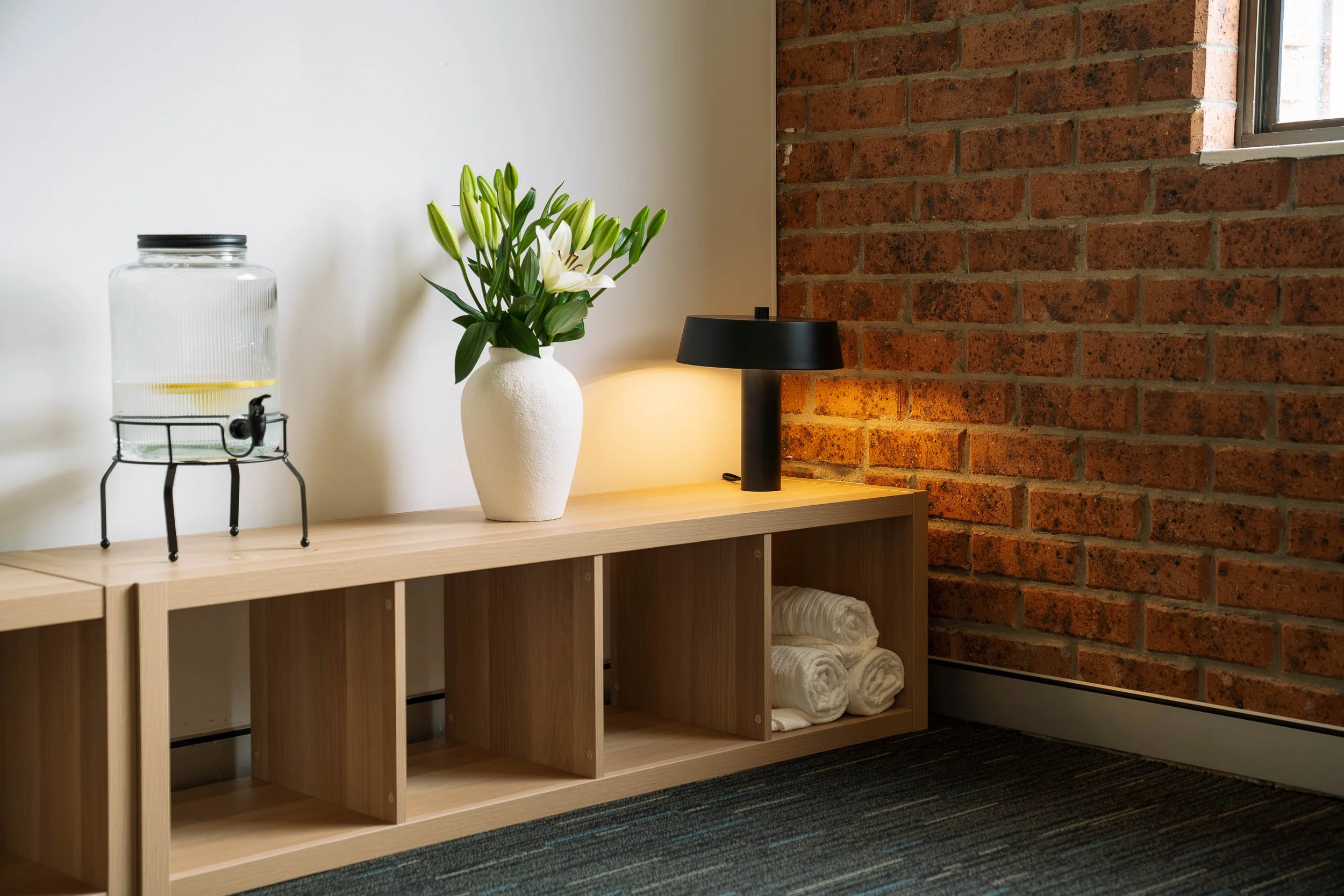 A light wooden shelf with open cubbies holding white towels, a white vase with white lilies, a black table lamp, and a glass water dispenser with a black lid and spigot, against a wall with exposed red brick on one side and plain white on the other.