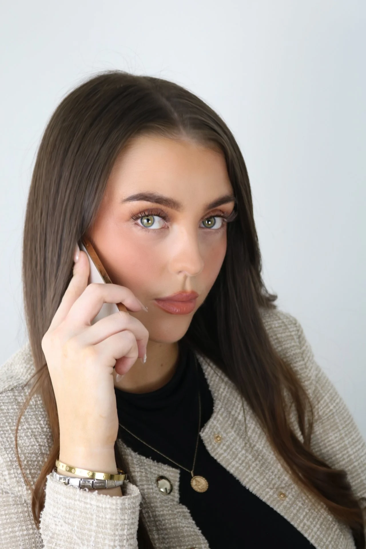 Women with long brown hair and green eyes talking on a smartphone, wearing a beige blazer and black top with gold jewelry.