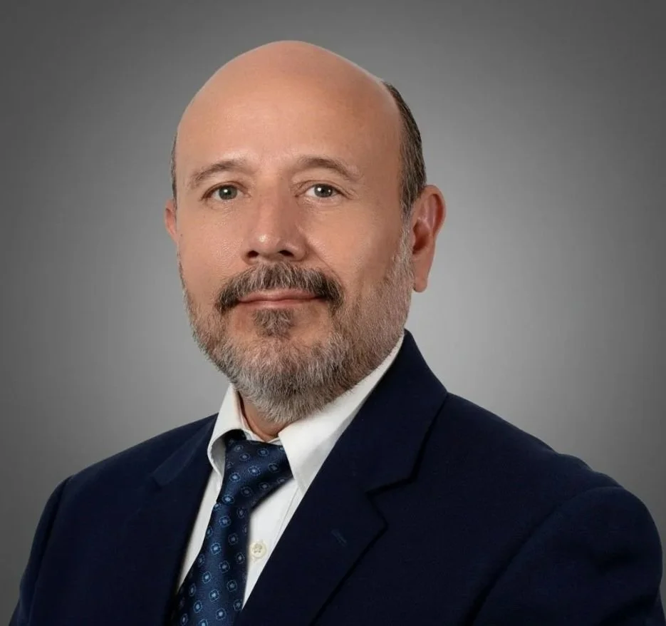 A middle-aged man with a bald head, beard, and mustache wearing a navy suit, white shirt, and patterned blue tie against a gray background.