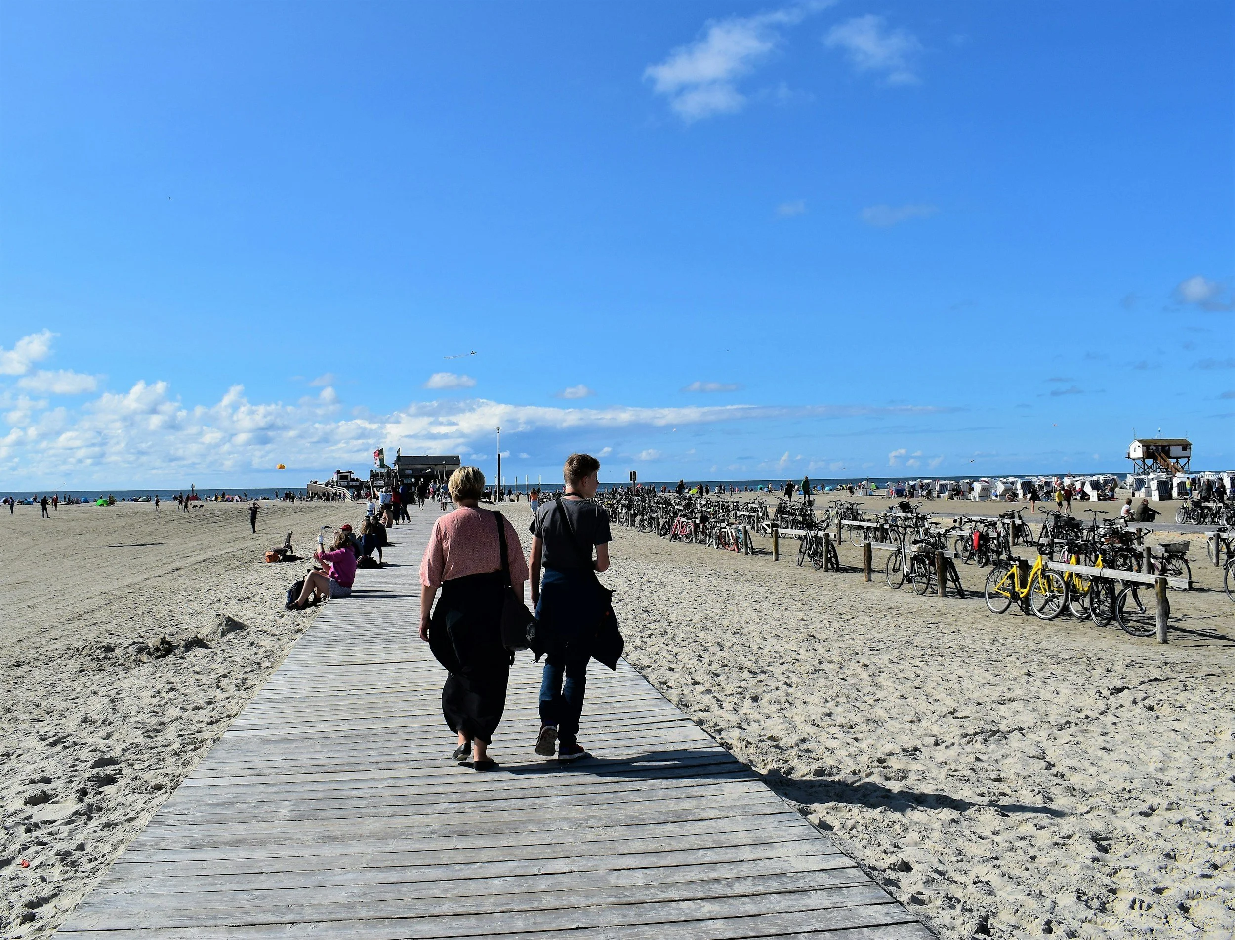 People walking on a wooden boardwalk on a sandy beach with bicycles parked along the side, under a blue sky with scattered clouds.