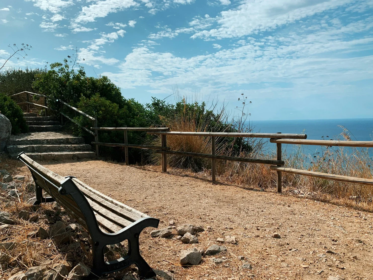 A wooden bench on a dirt path overlooking the ocean, with a railing, shrubbery, steps, and a cloudy blue sky.