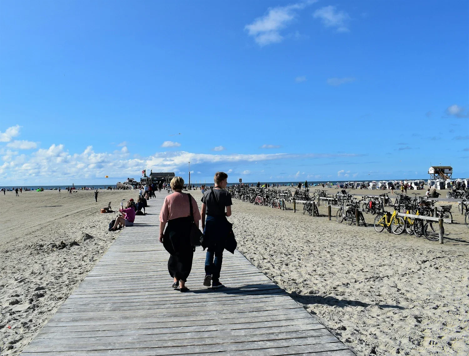 People walking on a wooden pathway along a sandy beach with rows of parked bicycles and a clear blue sky.