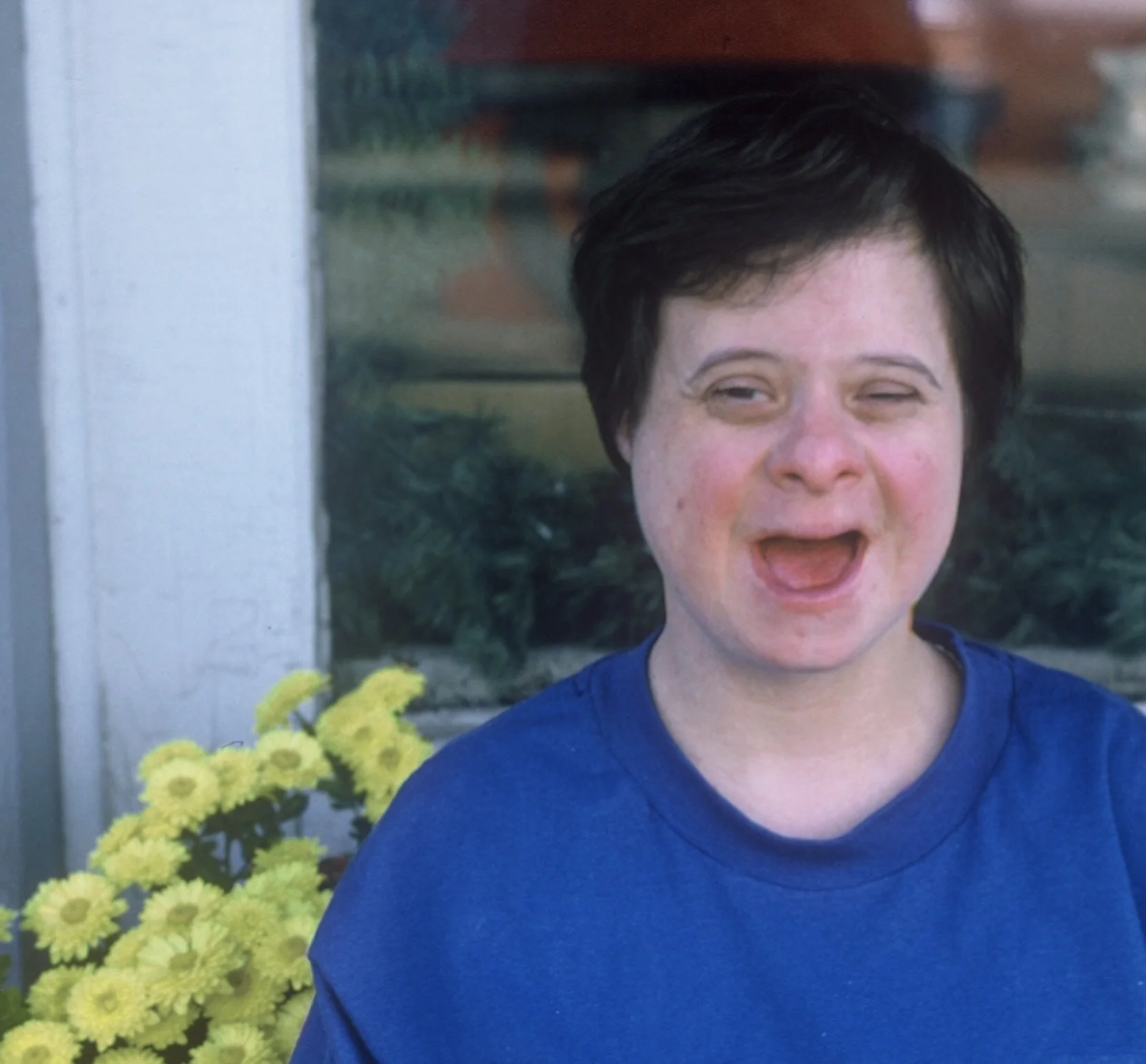 A young woman with short dark hair, wearing a blue shirt, standing outside near a window with yellow flowers behind her. She appears to be mid-sentence with an expressive face.