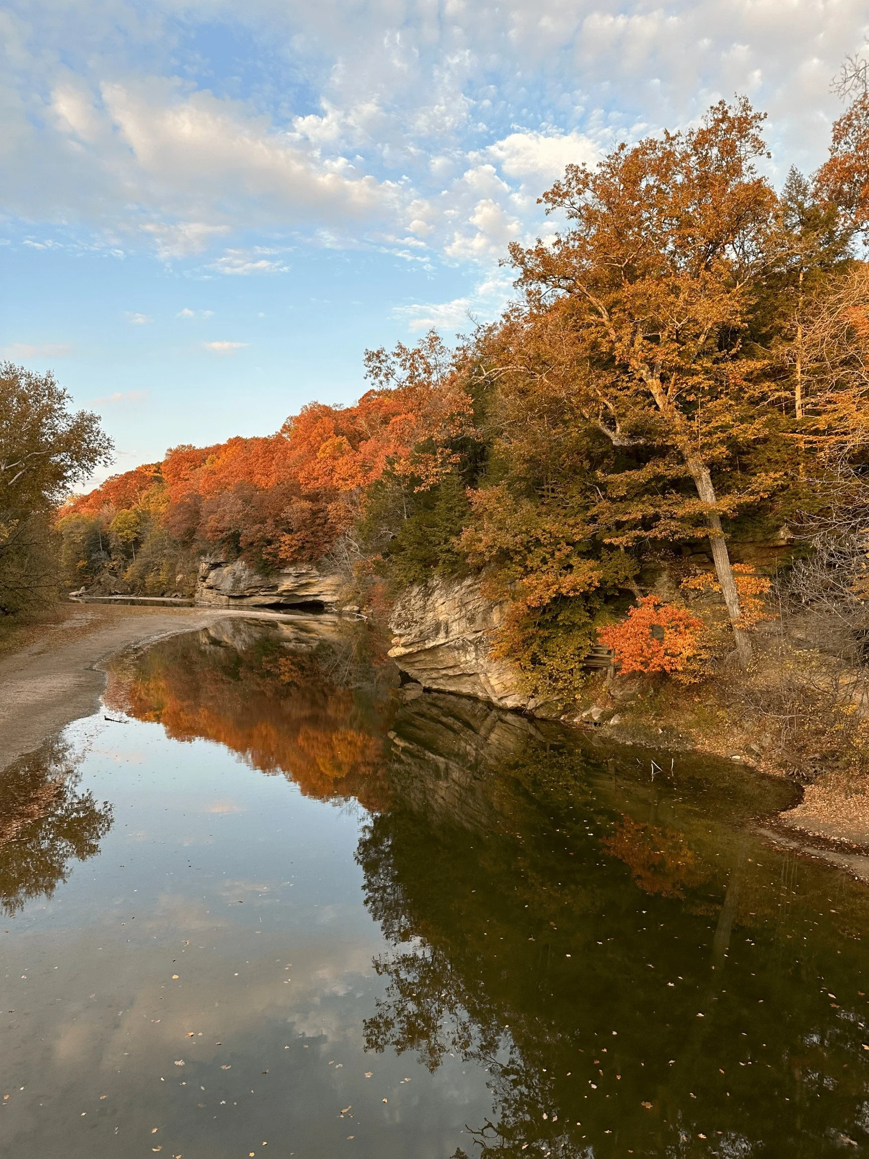 Autumn landscape with a reflective river, orange and green trees on rocky cliffs, and a partly cloudy sky.