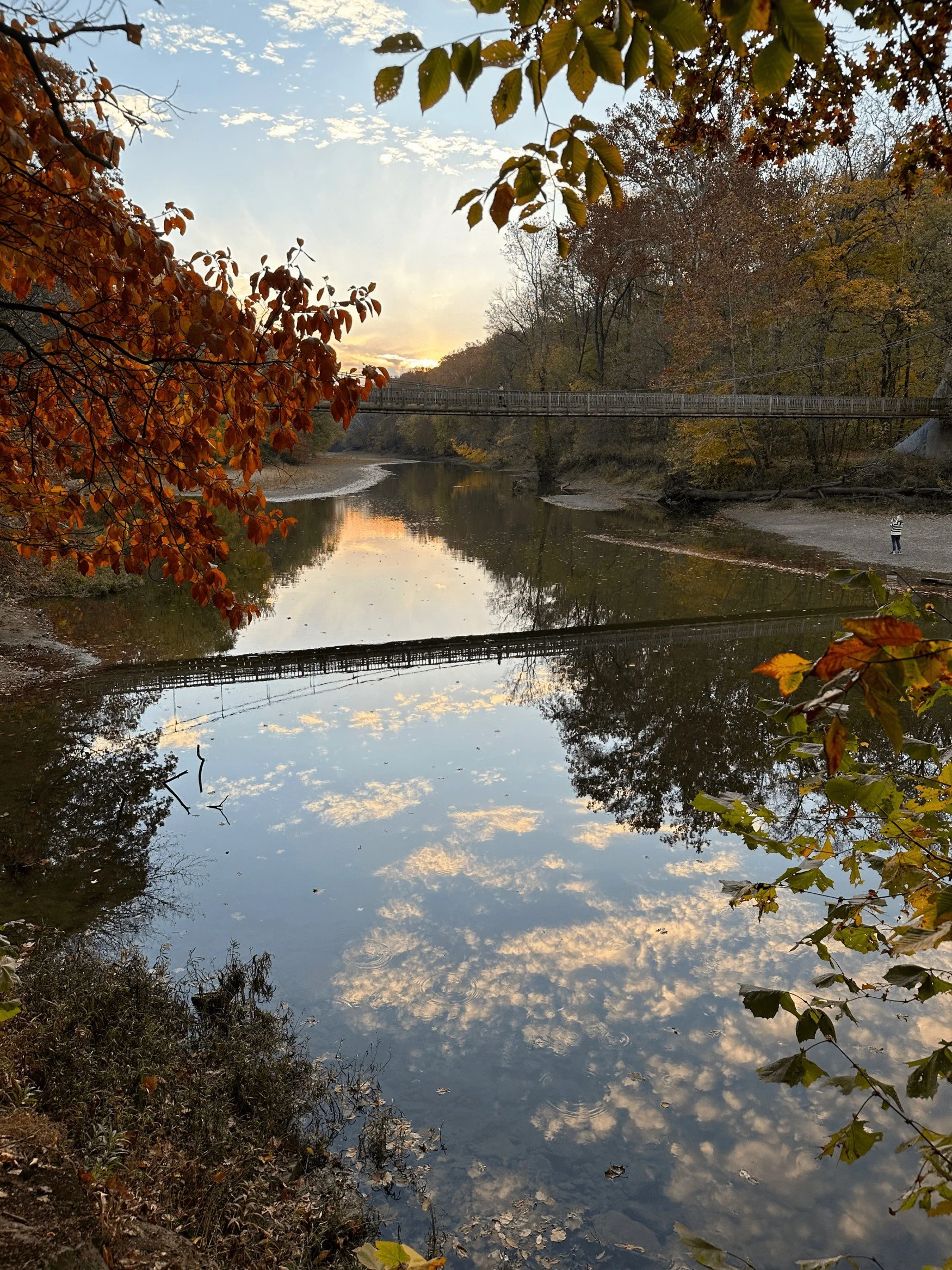 A calm river at sunset surrounded by fall foliage, with orange, yellow, and green leaves, and a bridge crossing over the river. Reflections of the sky and clouds are visible in the water.
