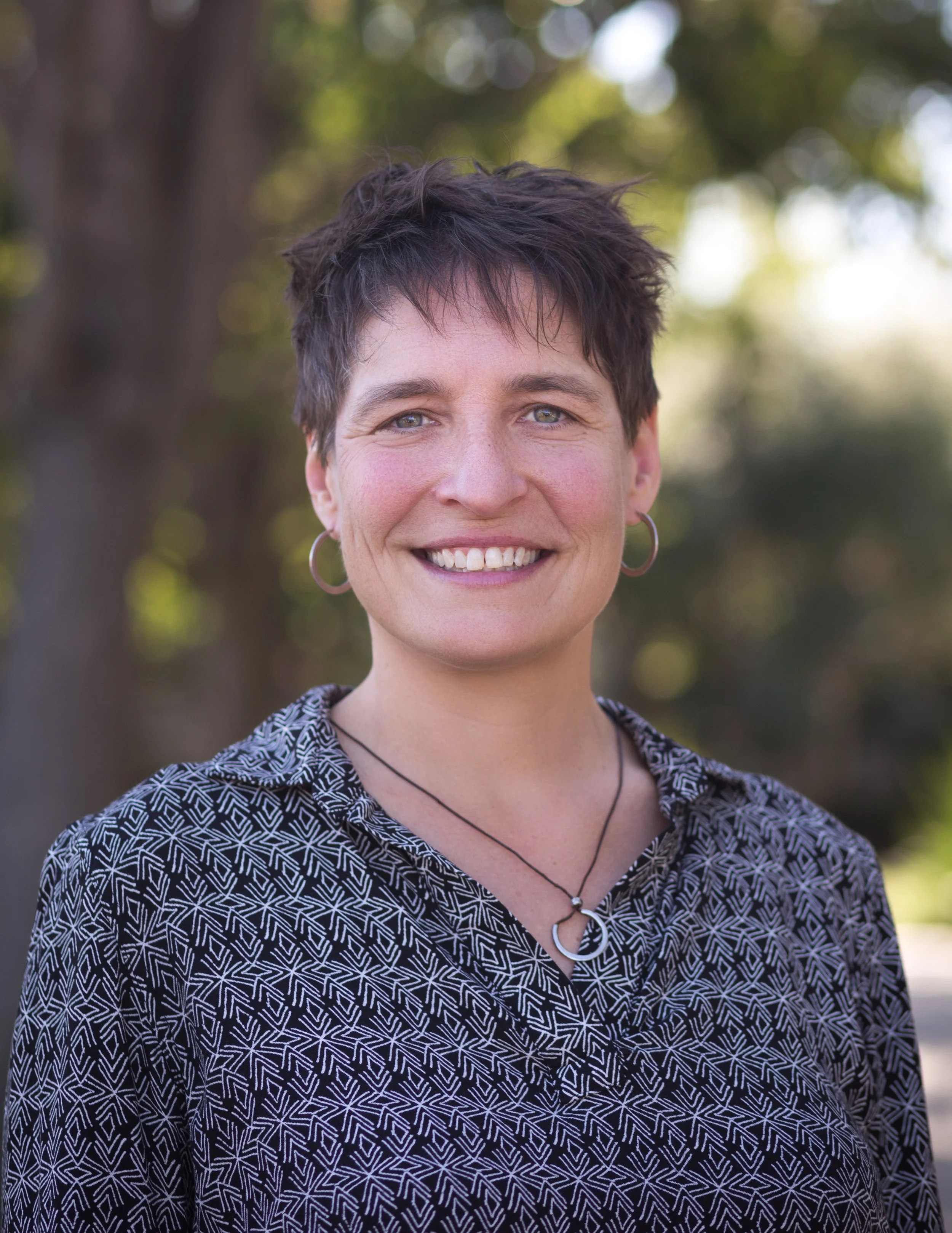 A woman with short dark hair and hoop earrings smiling outdoors, wearing a patterned shirt and a necklace with a moon pendant.