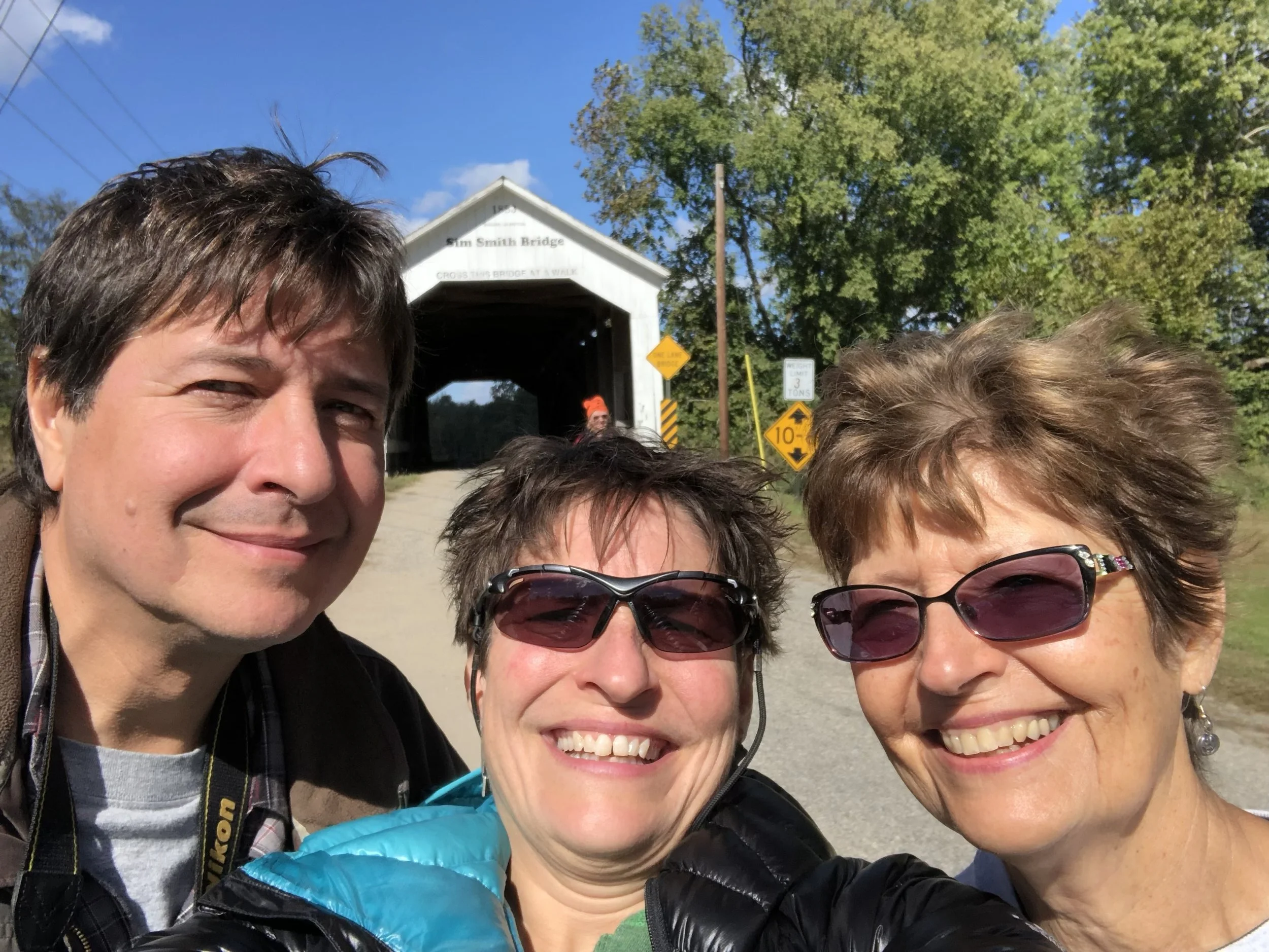 Three people taking a selfie in front of the Slim Smith Bridge on a sunny day, smiling, with trees and road signs in the background.