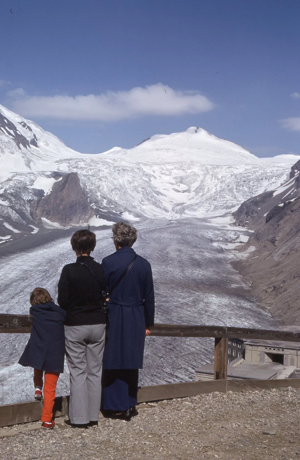 Three people, two women and one child, stand on a viewpoint overlooking a glacier, snow-covered mountains, and a blue sky with some clouds in the background.