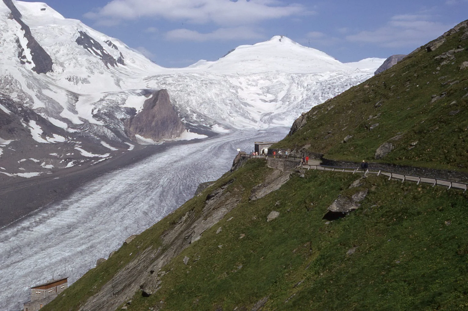 Scenic mountain landscape with snow-capped peaks, a glacier, and a winding trail with visitors walking along it.