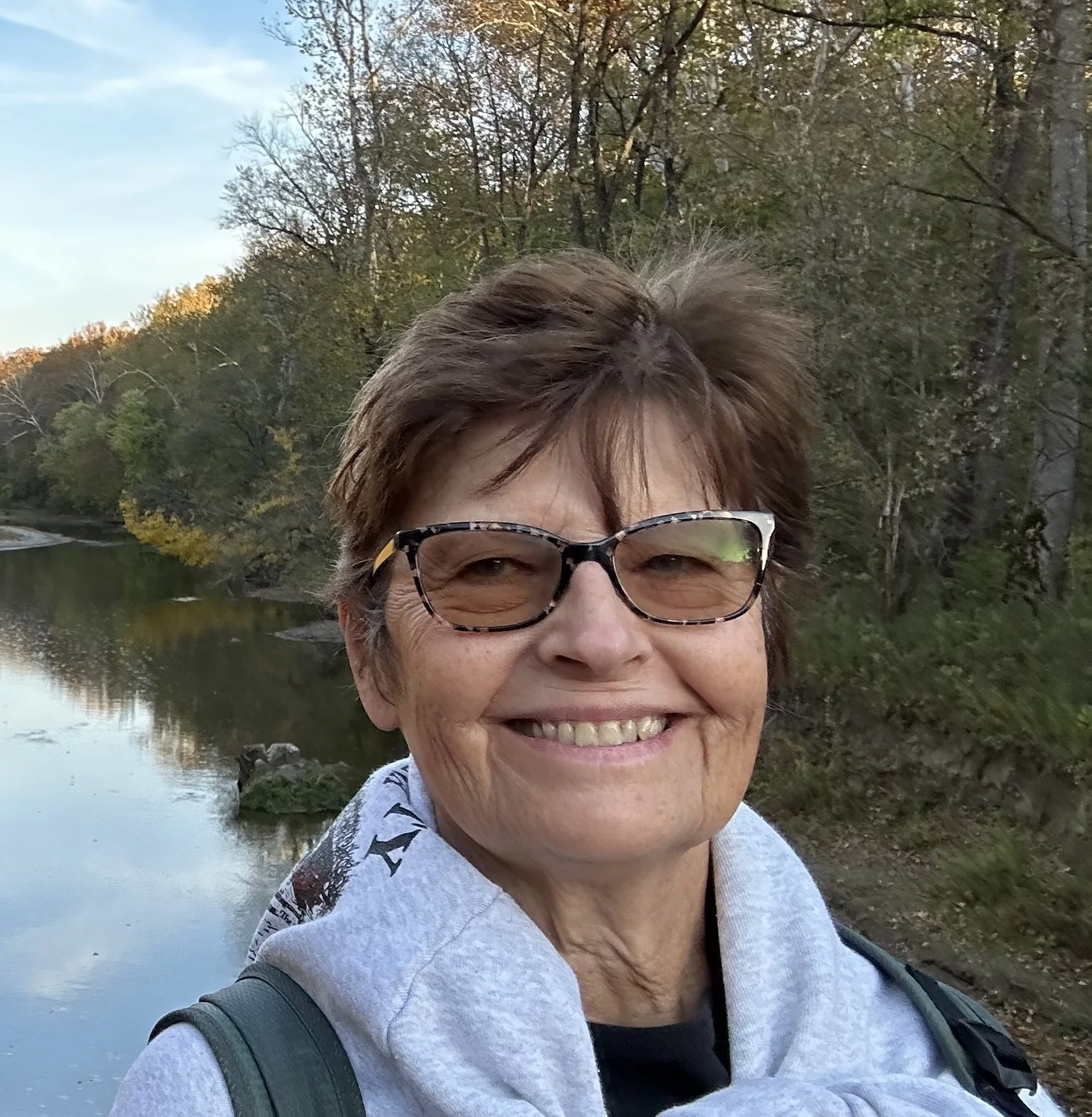 Smiling woman wearing glasses and a gray hoodie, standing outdoors near a body of water with trees in the background during what appears to be early evening.