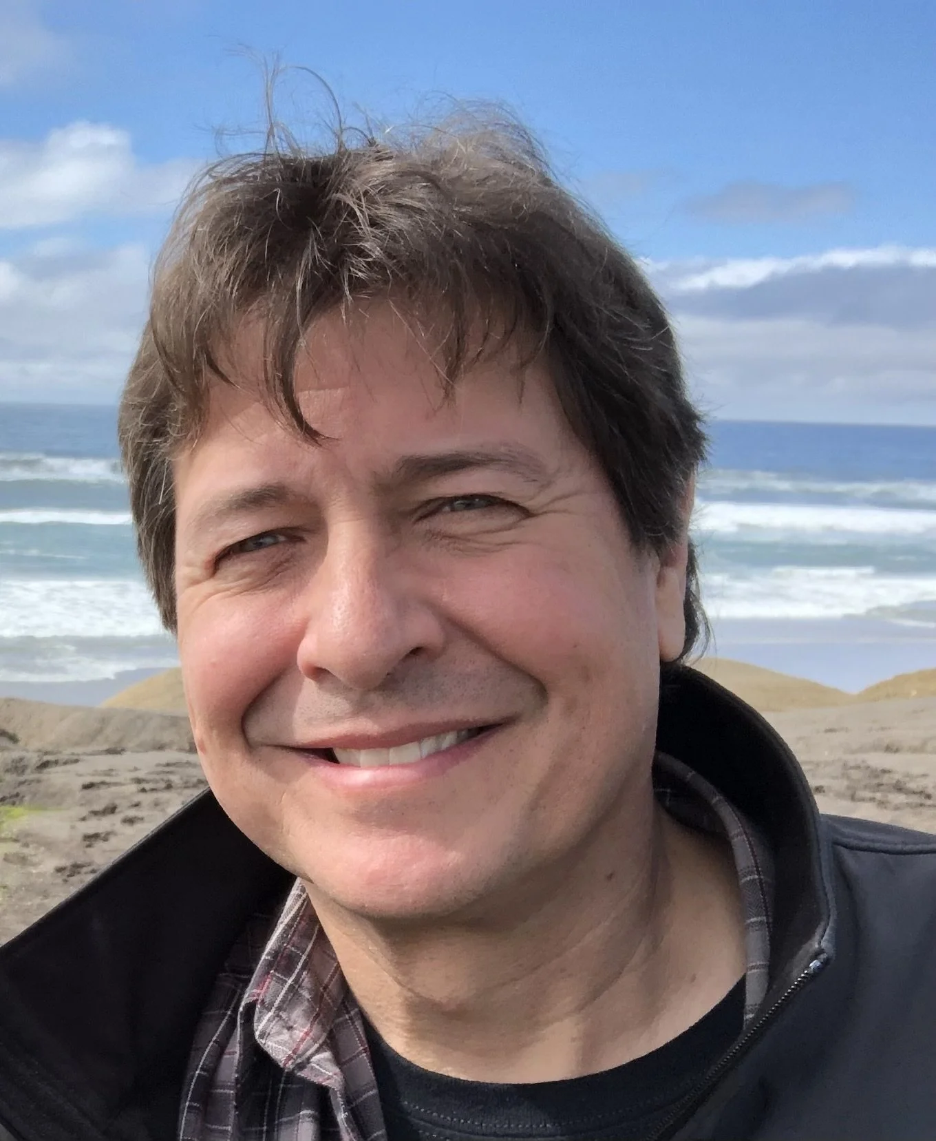 A smiling man with tousled brown hair outdoors near the ocean, with a sandy beach and cloudy sky in the background.