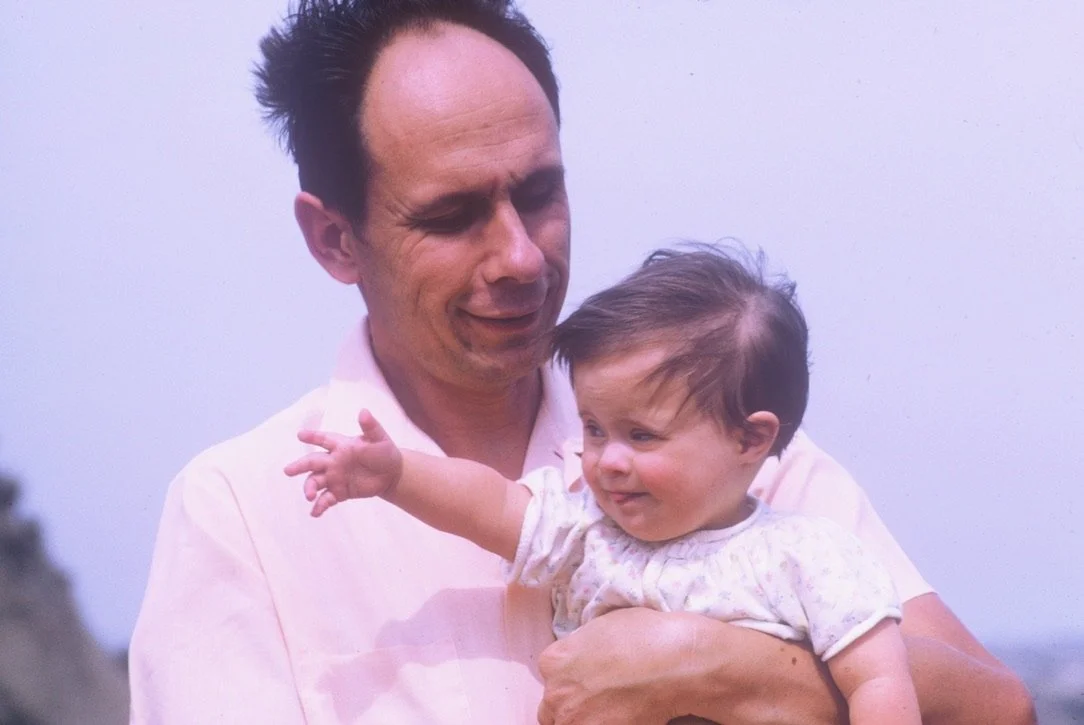 A man holding a smiling baby girl outdoors, both wearing light-colored clothing.