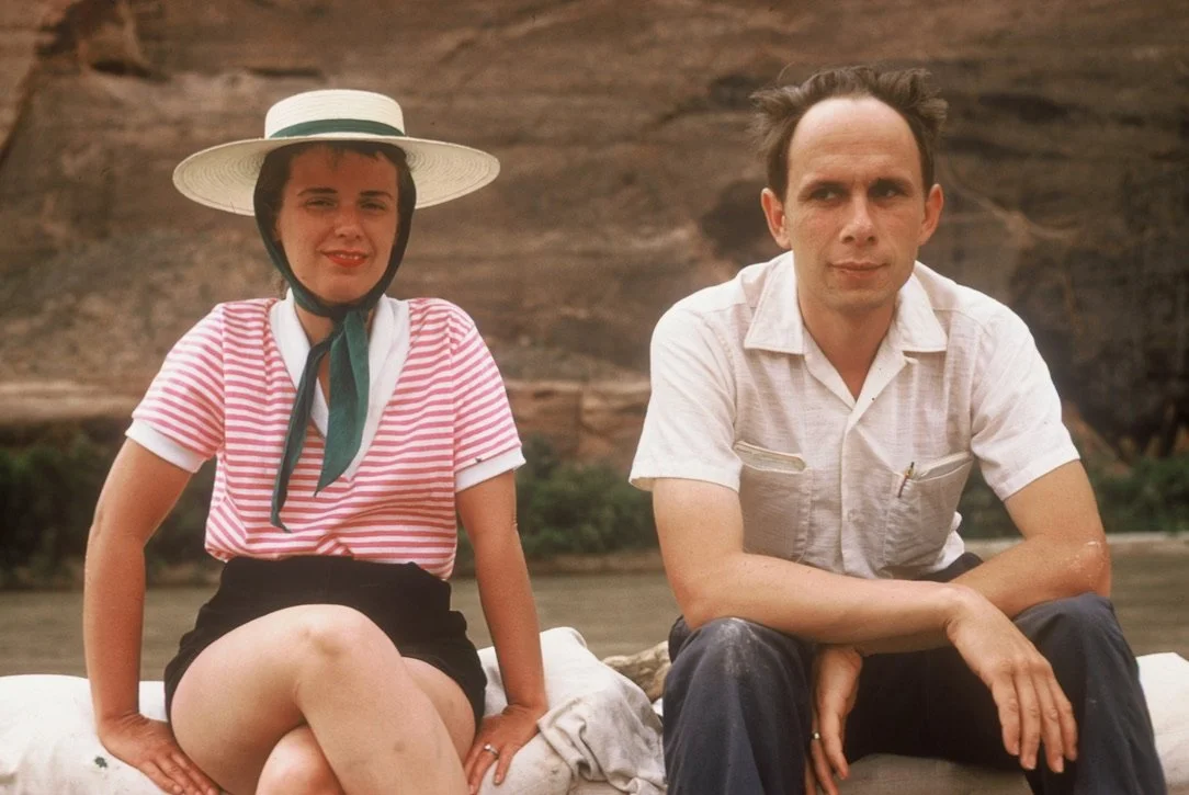 A woman wearing a wide-brimmed hat, striped shirt, and black shorts sitting beside a man in a white short-sleeved shirt, both outdoors in front of a rock formation.