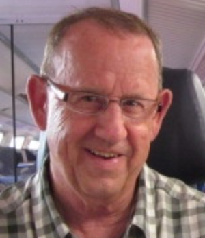 A smiling man with glasses and short hair, wearing a checkered shirt, sitting inside an airplane.
