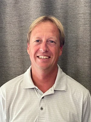 A smiling man with light-colored hair wearing a light gray polo shirt, standing in front of a gray curtain background.