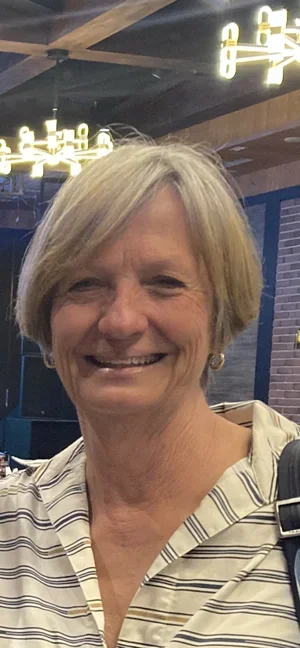 Smiling middle-aged woman with short blonde hair in a striped shirt, indoors with modern lighting fixtures.