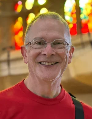 A smiling man with glasses and short hair wearing a red shirt, standing inside a church with colorful stained glass windows in the background.
