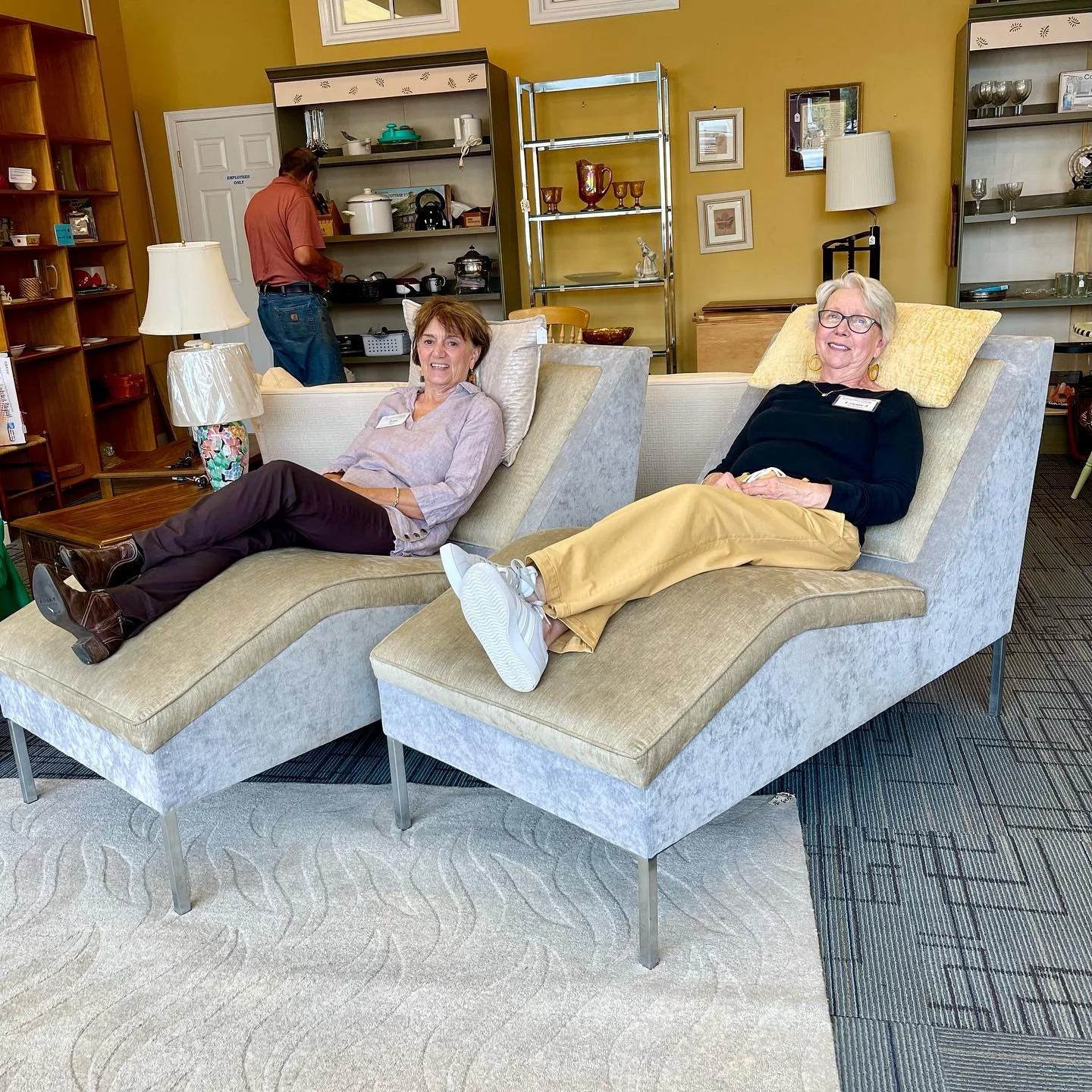 Two older women relaxing on beige chaise lounges in a furniture store. The woman on the left is wearing a light-colored shirt and dark pants, and the woman on the right is in a black top and yellow pants. A man in the background is browsing shelves.