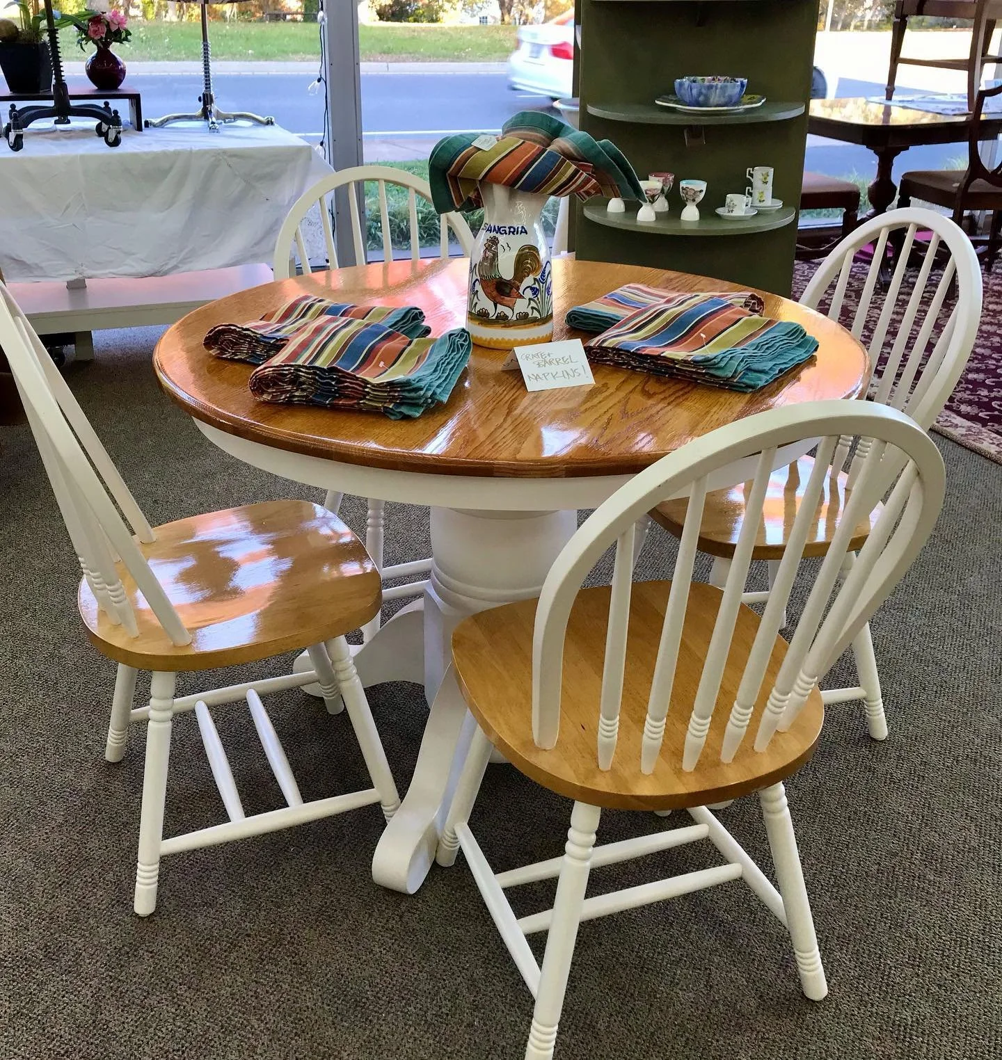 Round wooden dining table with four matching chairs, two with white spindled backs and two with natural wood seats, displayed indoors. The table is decorated with a ceramic vase labeled 'Sangria' containing striped napkins, and each place setting has