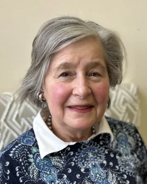 Close-up portrait of an elderly woman with short gray hair, smiling softly, wearing a white collared shirt and a patterned blue jacket.