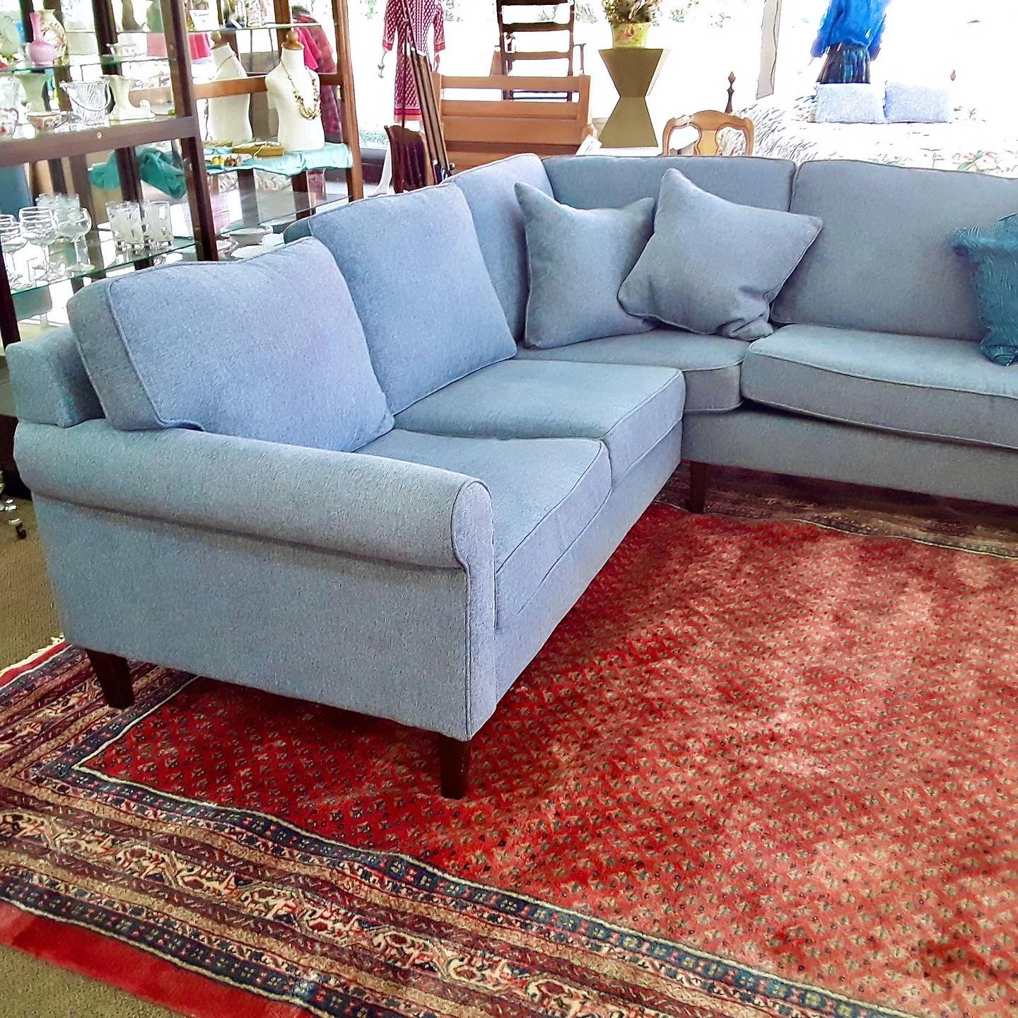 A light blue sofa with matching pillows, placed on a red patterned Persian-style rug. The background features display shelves with various household items and glassware, and some wooden furniture.
