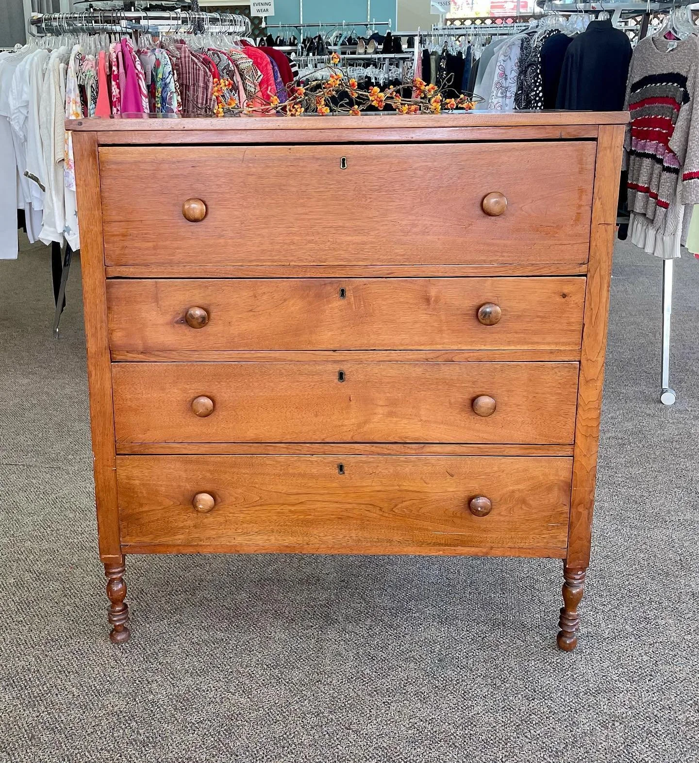 A wooden dresser with four drawers and round knobs, located in a clothing store with racks of clothes in the background.