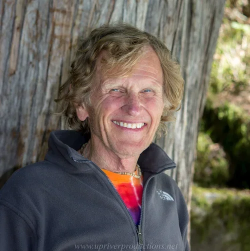 Smiling man with curly blonde hair wearing a black North Face jacket and an orange tie-dye shirt outdoors in front of a wooden fence.