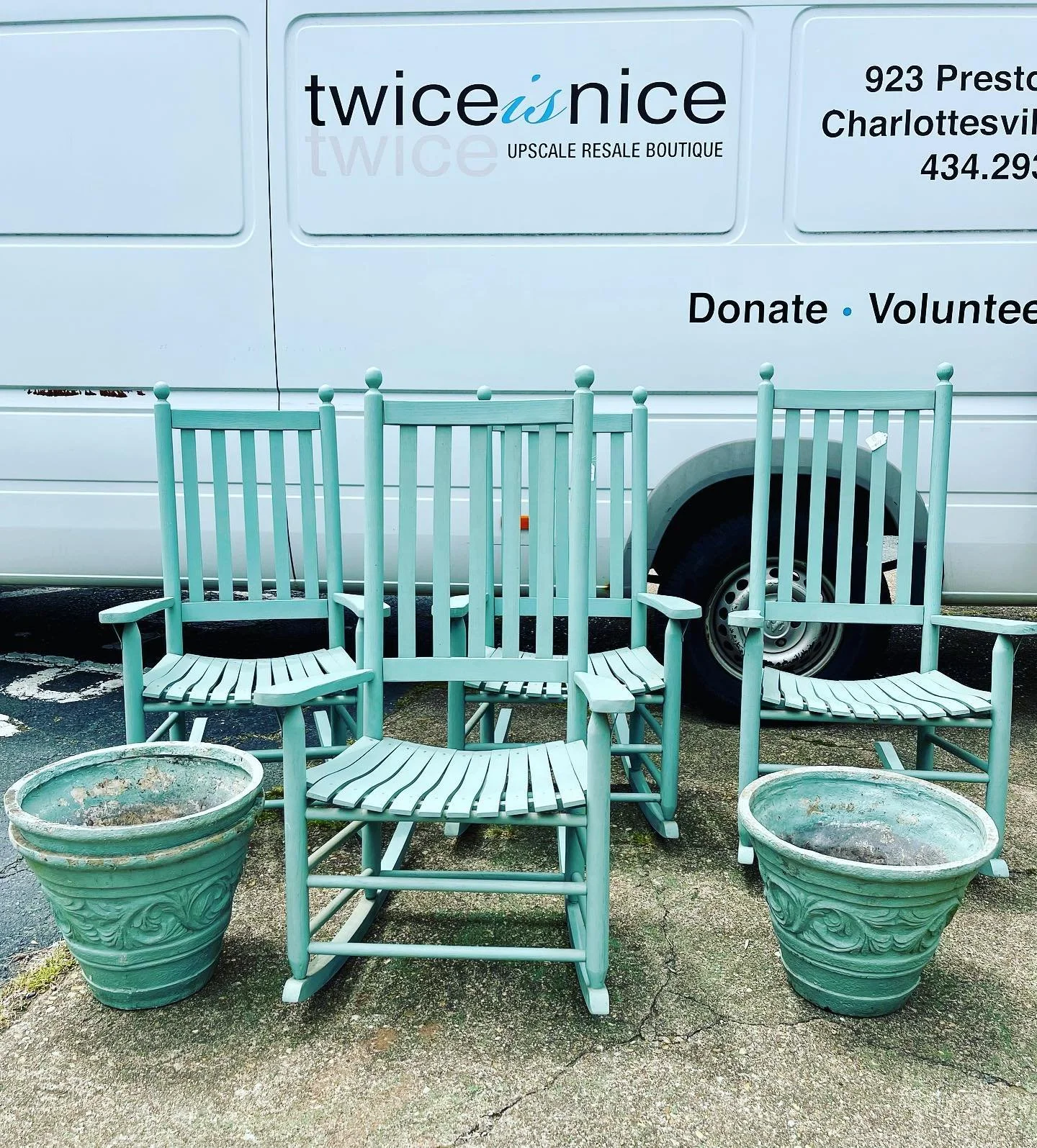 Four teal wooden chairs with armrests and slatted seats and backs, two empty decorative planters, are placed outdoors on a paved surface, with a white van in the background displaying a sign for "twice is nice Upscale Resale Boutique".