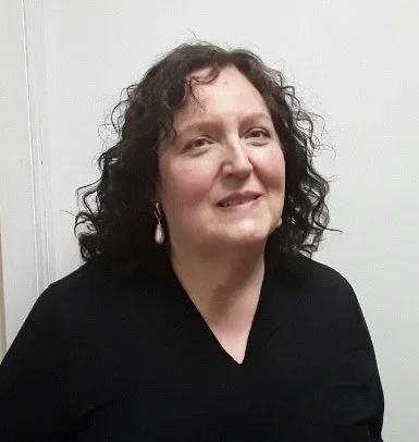 A woman with curly brown hair wearing a black top and earrings standing against a white wall.