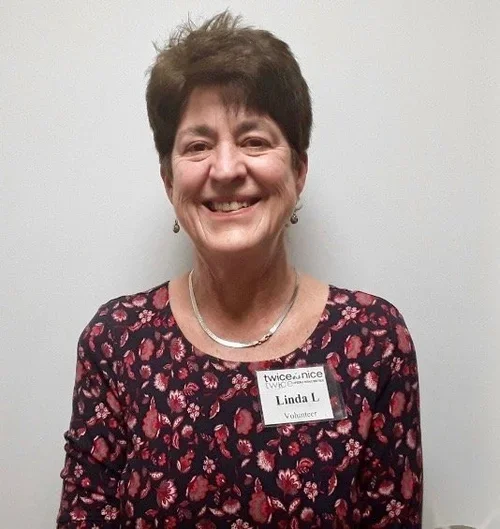 A smiling woman with short dark hair wearing a floral dress and a name tag that says 'Linda L, Volunteer'.