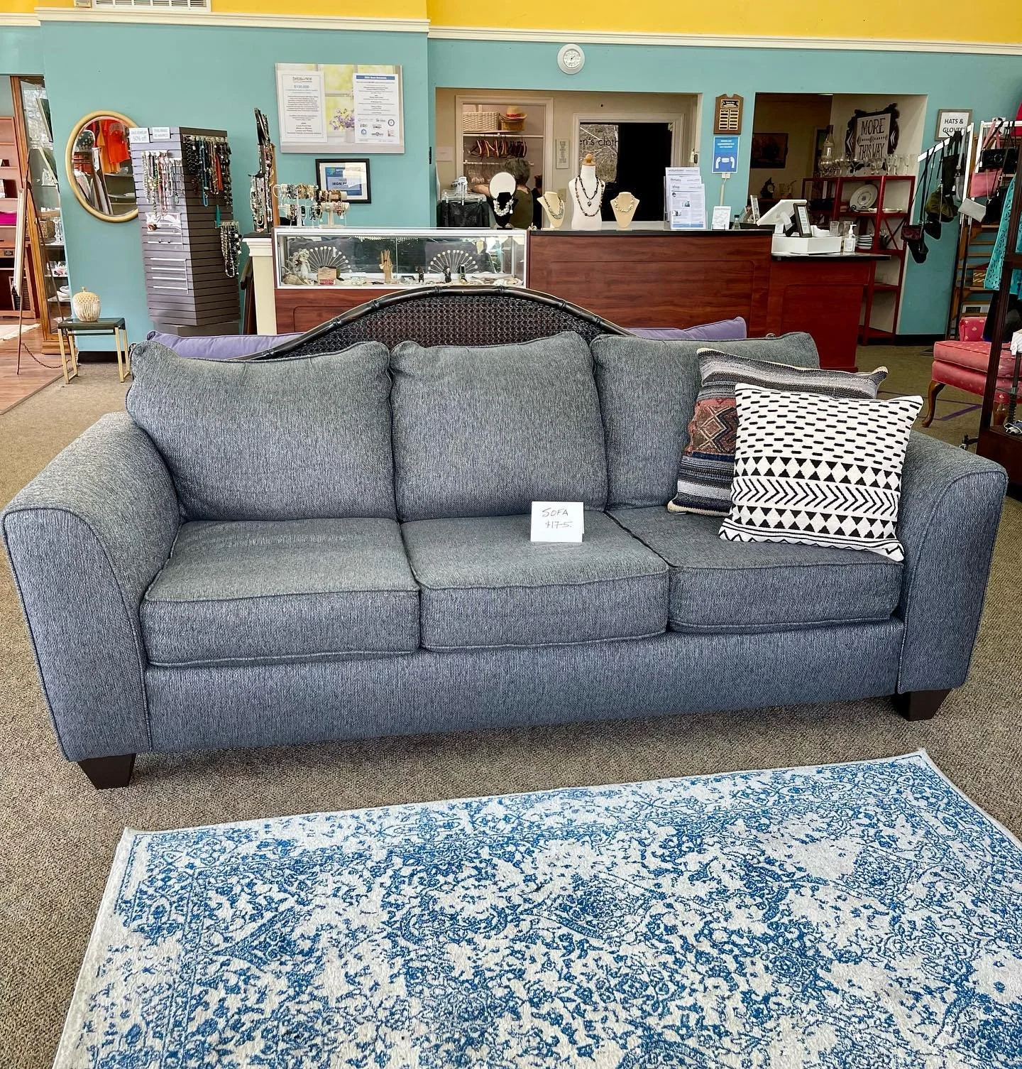 Gray three-seat sofa with patterned pillows on a blue and white rug in a furniture store.