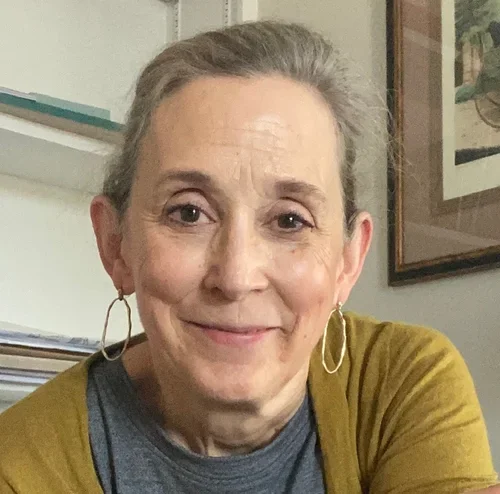 A woman with short gray hair, wearing hoop earrings, a blue shirt, and a yellow cardigan, smiling indoors with framed pictures and shelves in the background.