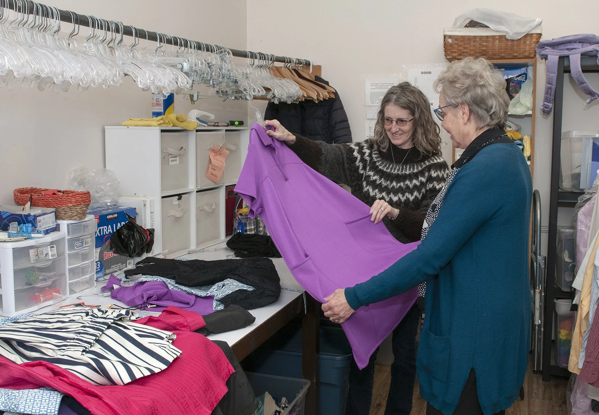 Two women shopping for clothes at a thrift store, holding a bright purple garment and smiling.