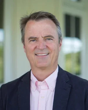 A smiling man in a pink shirt and navy blazer standing outside a house.