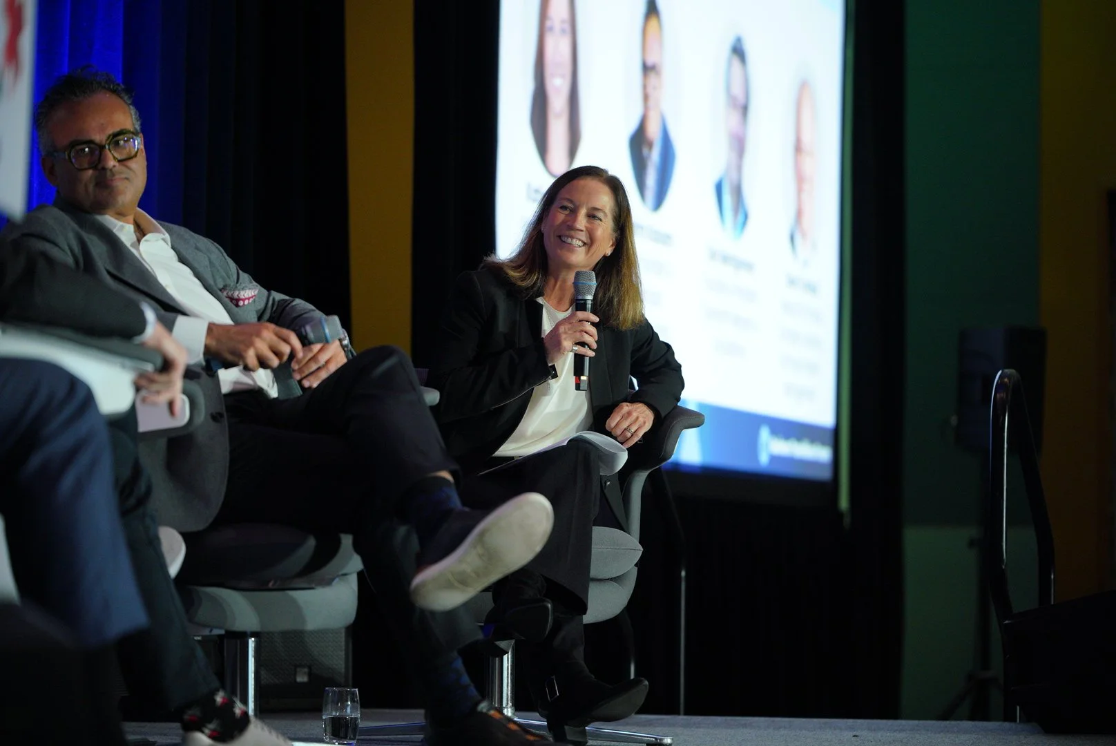 A woman holding a microphone, sitting on a panel during a conference or discussion, with a large screen displaying her image and other panelists in the background.