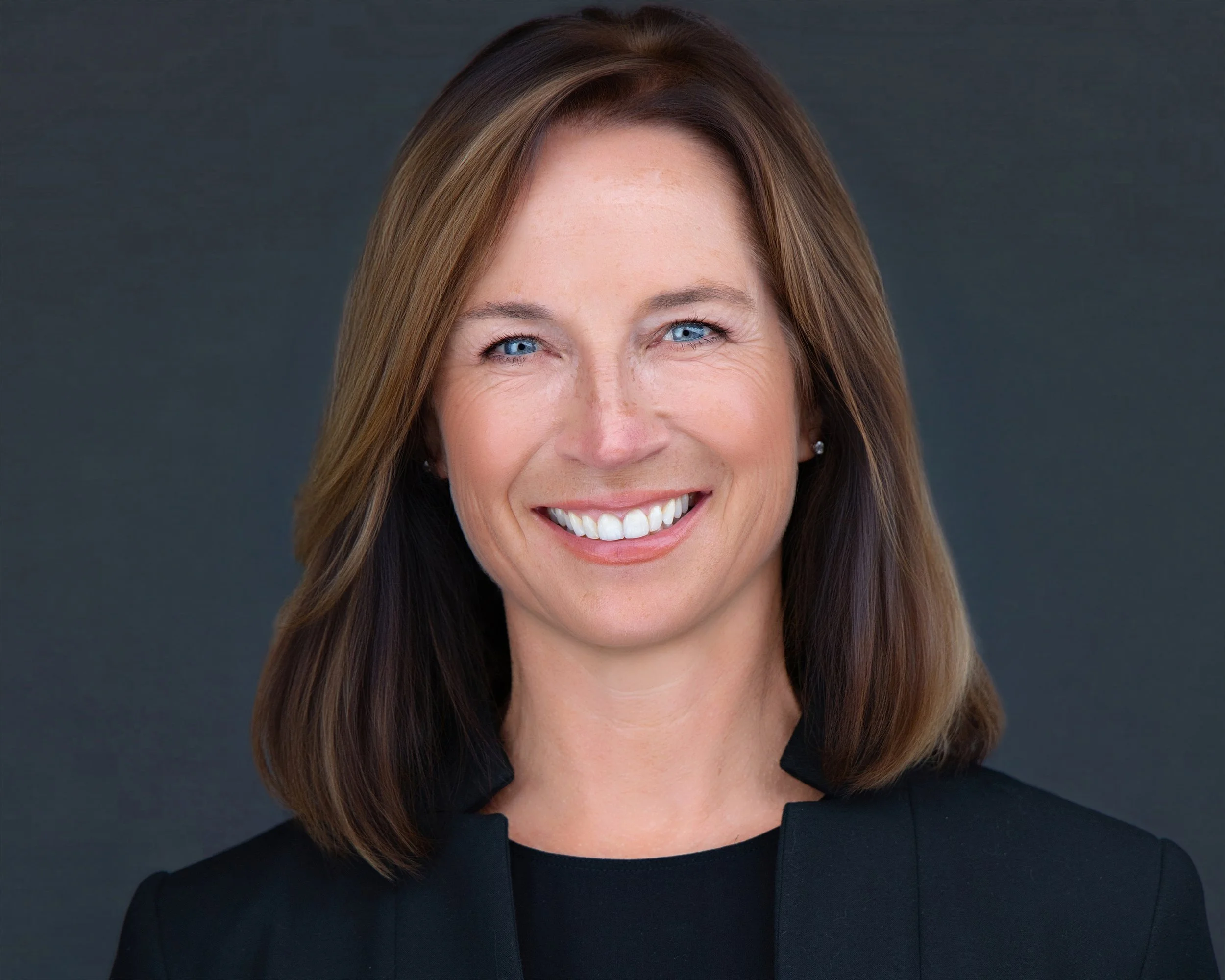 Headshot of a woman with shoulder-length brown hair, blue eyes, wearing a black blazer, smiling against a dark background.