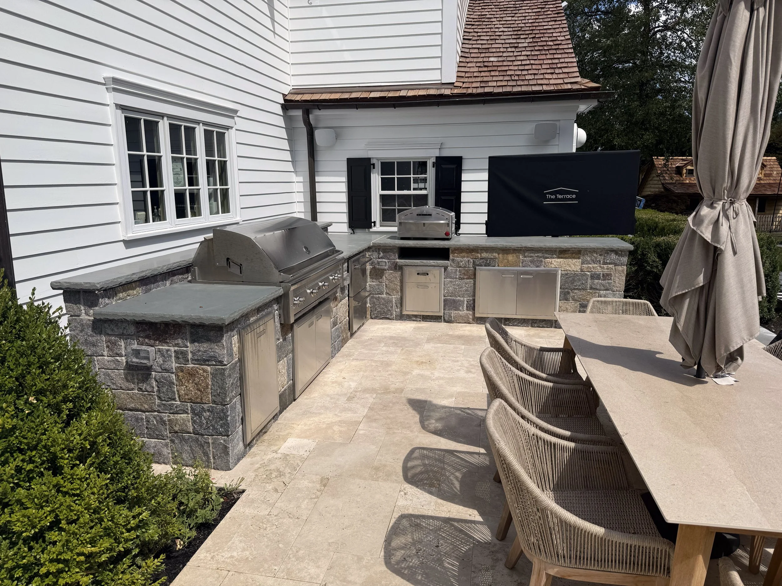 Side view of a masonry kitchen with bluestone countertops.  Travertine deck with dining table.  54" Lynx grill with Pizza Oven and outdoor TV