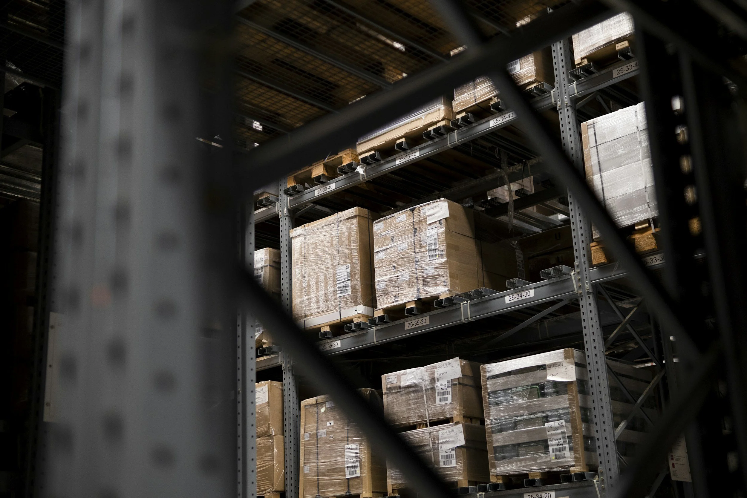 View of storage shelves filled with cardboard boxes wrapped in plastic in a warehouse, seen through a black metal rail in the foreground.