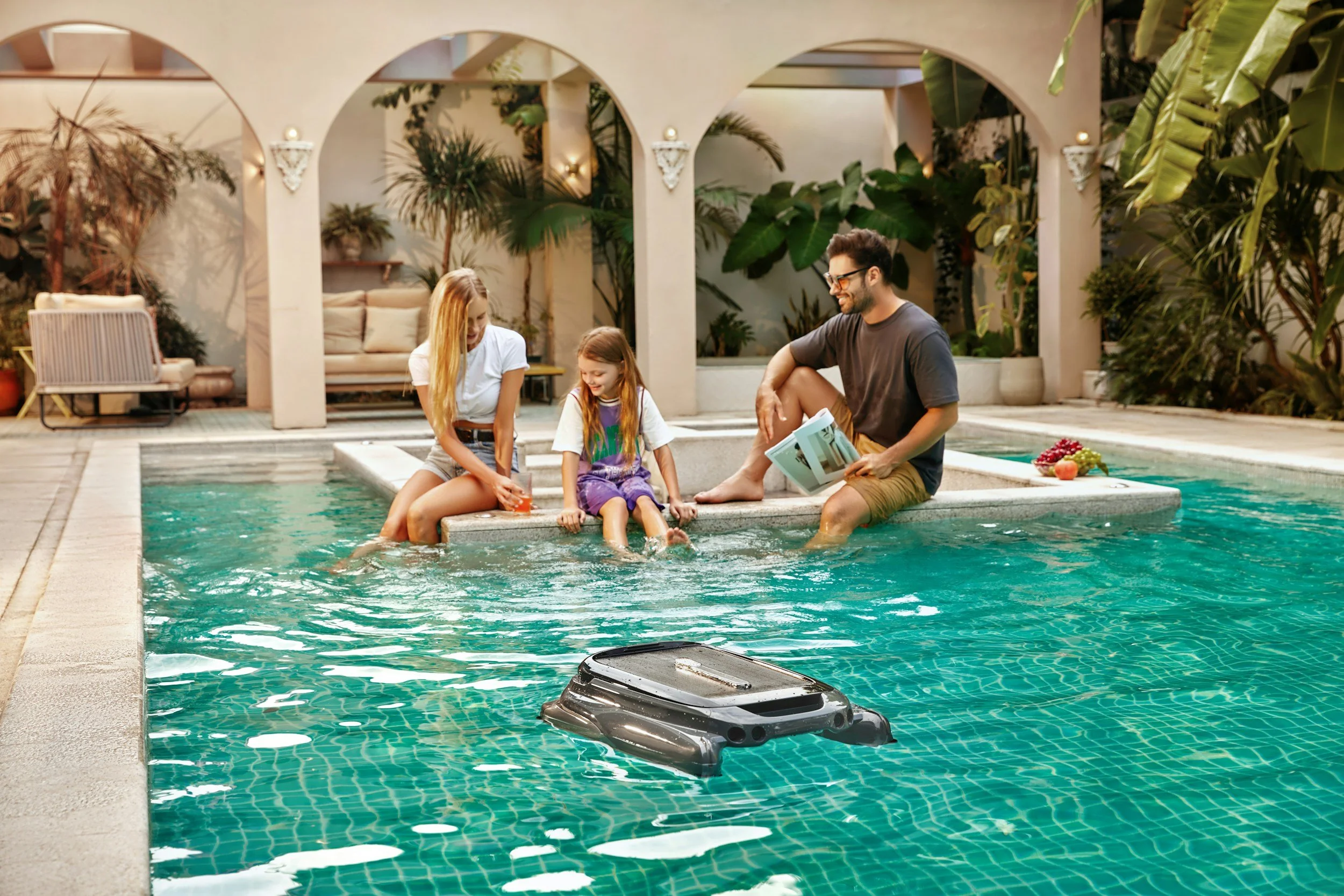 A family of four sitting by a swimming pool, with two children and two adults, in a lush, tropical outdoor setting.